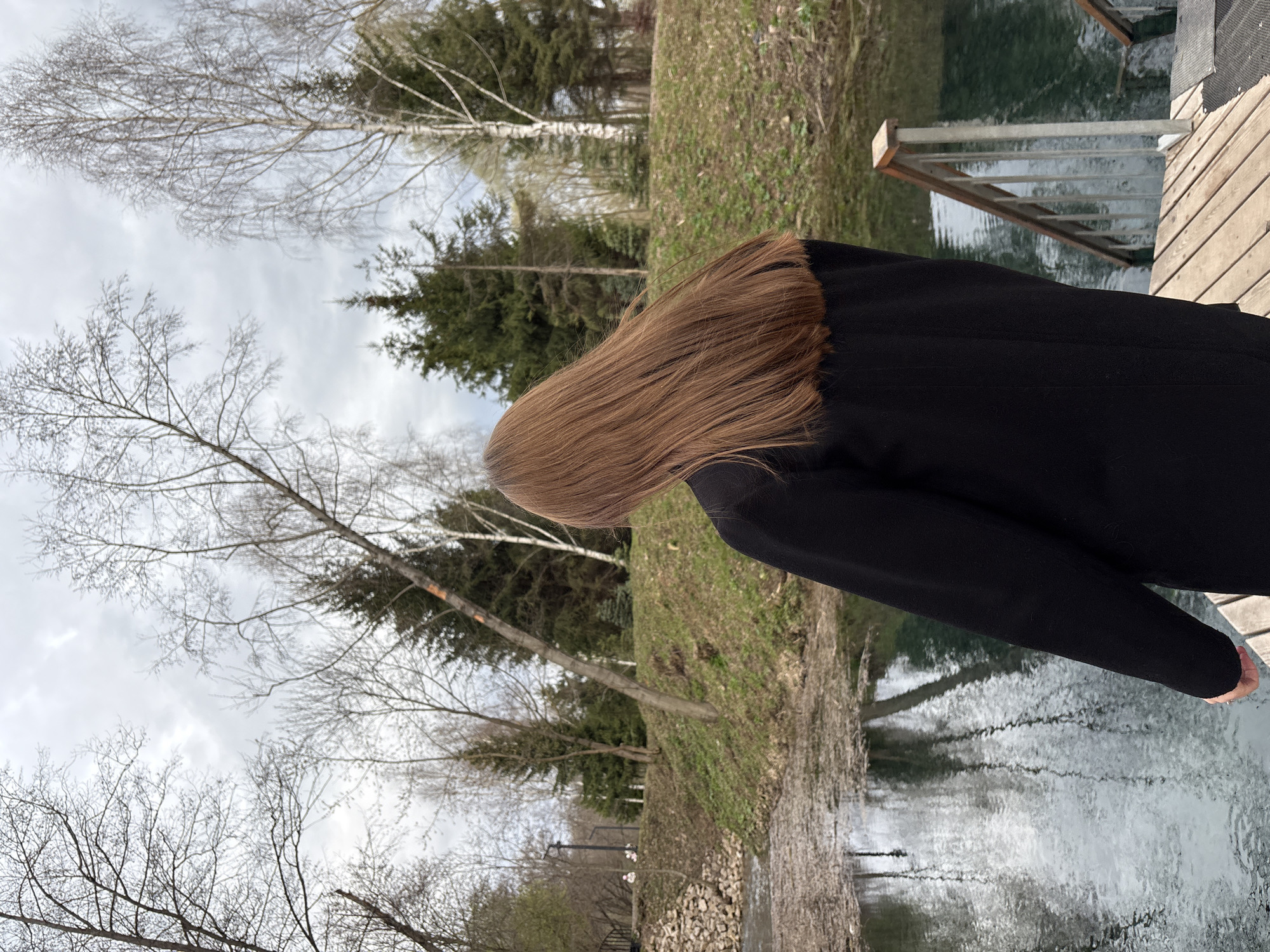 The image, oriented upright, depicts an outdoor scene in Sdt Shcherbakovka, Russia, featuring a person standing by a body of water.
In the foreground, the back of a person is visible, characterized by long, light brown hair flowing down their back. They are wearing a dark, possibly black, coat or jacket, and their right hand is resting on a wooden surface.
The person appears to be standing on a wooden dock or platform that borders a calm body of water, likely a river or lake. A metal railing structure is visible extending from the dock into the water. The water's surface reflects the cloudy sky and surrounding landscape, showing gentle ripples.
Beyond the water and behind the person, there is a natural bank covered with grass and sparse, dry vegetation. A line of trees forms the backdrop, consisting of a mix of deciduous trees with bare branches, indicative of late winter or early spring, and coniferous trees with dark green foliage. The sky above is overcast with light grey clouds.
The scene suggests a quiet, contemplative moment. The person is not engaged in any dynamic activity, appearing to be observing the environment. The light is diffused, characteristic of an overcast daytime. No visible text is present in the image.