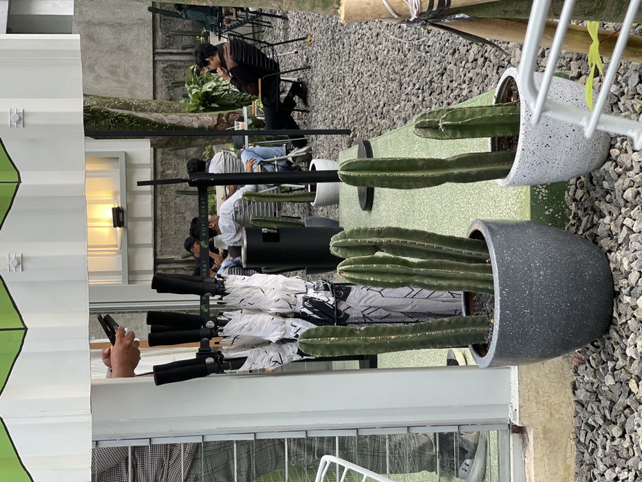 The image depicts an outdoor cafe area, identified as a "Kopi Sobat" establishment, located in Bogor, Indonesia. The ground is covered uniformly with light grey gravel.
In the foreground, two large pots containing tall, green columnar cacti are prominently positioned. The pot on the left is dark grey and speckled, holding multiple cacti, while the pot on the right is white and speckled, containing two cacti and partially obscured by a white metal railing.
The midground features a white building structure with a green, geometric-patterned roof overhang. Beneath this overhang, a stand holds numerous folded white umbrellas with black handles and black-and-white patterned fabric. Above the umbrellas, a person's hands are visible, holding a black smartphone that appears to be in a folded or flip position. To the left of the umbrella stand, a partial reflection of a person wearing plaid pants and dark shoes is visible in a glass surface.
The background reveals an outdoor seating area occupied by at least five individuals. Three people are seated at a table on the left, one wearing a light-colored hoodie and looking to the right. Two people are seated at a table on the right; one individual in a dark long-sleeved shirt and dark pants is leaning back in a black metal chair, with their right hand near their chin. Small, light-colored round tables and black metal chairs are distributed across the gravel. Additional potted cacti are interspersed within the seating area. A black sign displaying "KOPI SOBAT" in white text is visible above the seated patrons. Green foliage and trees are visible in the distant background against a grey wall, and a window on a white wall in the upper background emits a warm yellow light.