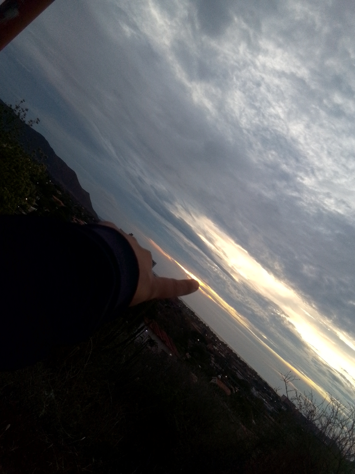 The image, taken at an oblique angle, shows a landscape at dusk. A dark mountain is visible on the left. The sky is filled with clouds, with a bright, horizontal band of light suggesting the setting sun. The lower portion of the image is dark, showing foliage and possibly the edge of a city. A person's arm and hand, pointing towards the horizon, are visible in the foreground. The city lights are visible on the water indicating the location is the bay area. Based on the details, the location is most likely Juan Griego, Venezuela, though this cannot be definitively confirmed from the image alone.