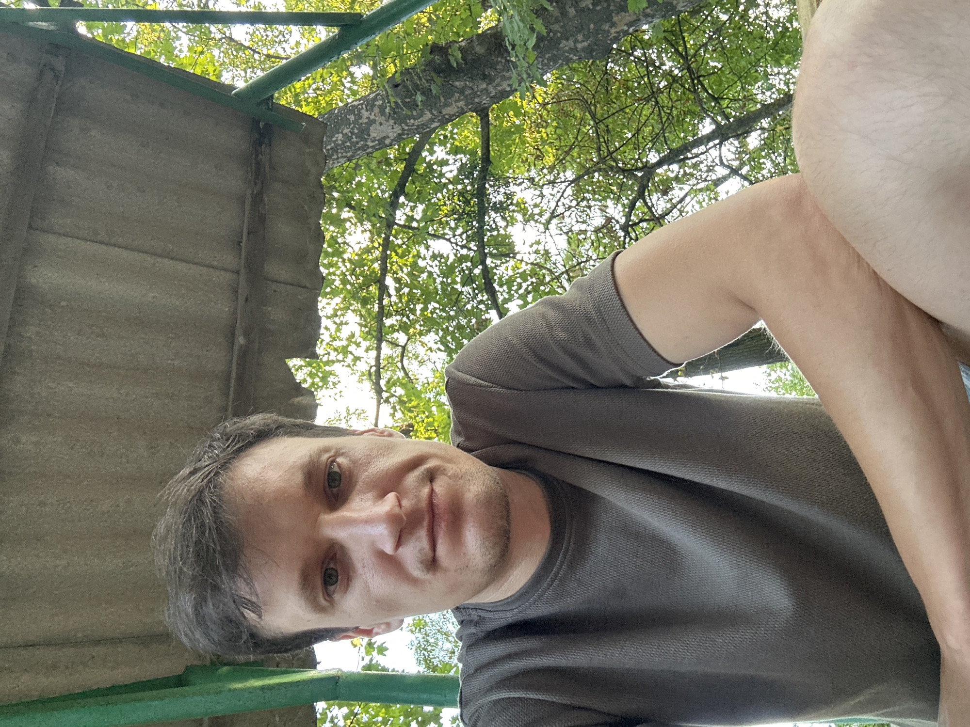 A man is pictured outdoors, possibly in a rural area or park in Makiv, Ukraine, as suggested by the surrounding trees and natural light. He is wearing a grey, short-sleeved t-shirt and is positioned at a low angle, looking directly at the camera. His expression is neutral. The background consists of lush green foliage and a tree trunk, indicating a natural setting. Above him, a rudimentary shelter made of corrugated material and supported by green metal poles is visible. The lighting suggests it is daytime, with sunlight filtering through the leaves. The man's arms are resting on his lap or a surface, and his posture is relaxed.
