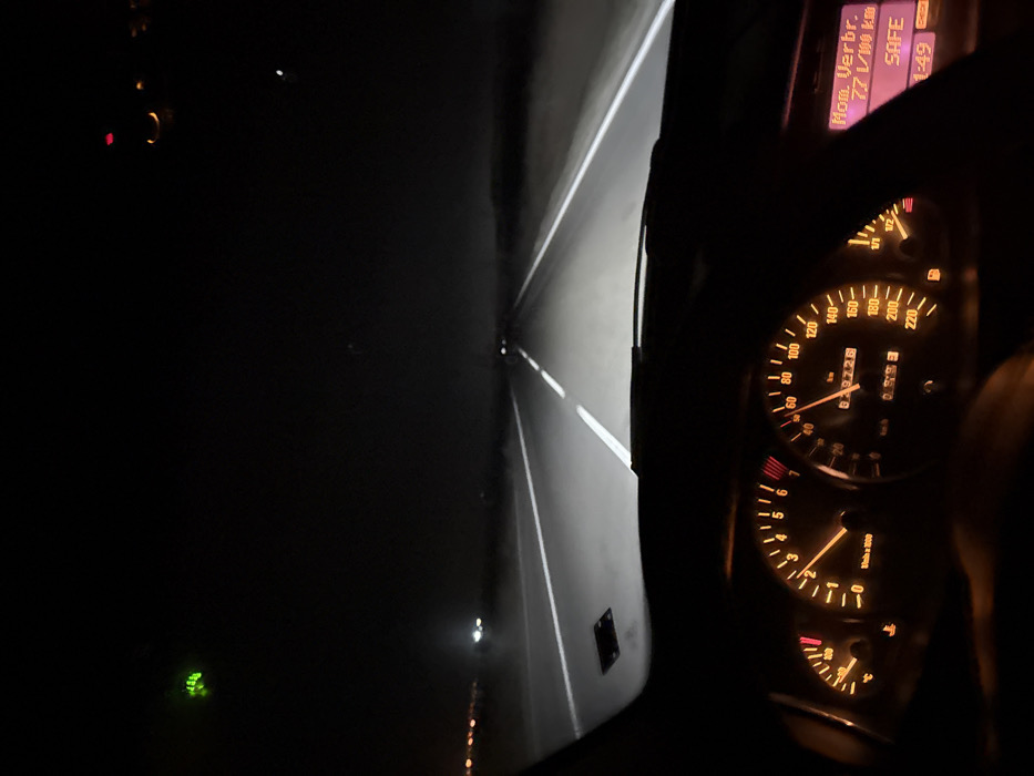The image presents a nighttime view from the driver's perspective inside a vehicle moving along a paved road in the city of Trapoklovo, Bulgaria. The road ahead is illuminated by the vehicle's headlights, revealing distinct white dashed lane markings and solid white shoulder lines. Distant lights are visible on the left horizon, suggesting an urban or industrial area. The vehicle's dashboard is illuminated with an orange glow, prominently displaying the speedometer indicating approximately 60 km/h and the tachometer at roughly 2000-2500 RPM. The odometer reads 829726 kilometers, and the trip meter shows 0099.3 kilometers. The fuel gauge indicates approximately one-quarter tank. A digital information display on the right side of the dashboard shows "Ком. Verb. 7.7 l/100 km", "SAFE", and "1:49". Multiple warning lights are active on the instrument panel. A green light source or reflection is present in the upper left of the frame. The presence of a driver operating the vehicle is inferred from the perspective and active dashboard instrumentation.