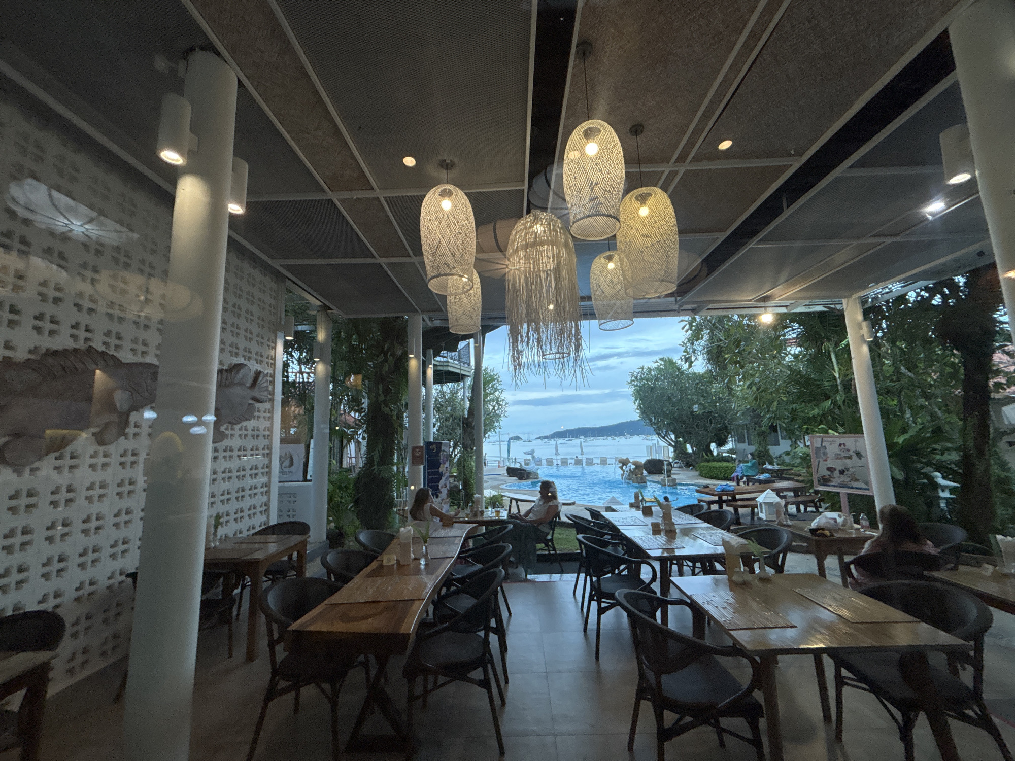 The image shows an interior view of a restaurant in Chalong, Thailand, with a large swimming pool and the ocean visible through a glass wall. The restaurant features wooden tables and chairs, with several patrons seated at the tables. Woven pendant lights hang from the ceiling, casting a warm glow. The atmosphere appears relaxed and inviting, with lush greenery surrounding the pool area.