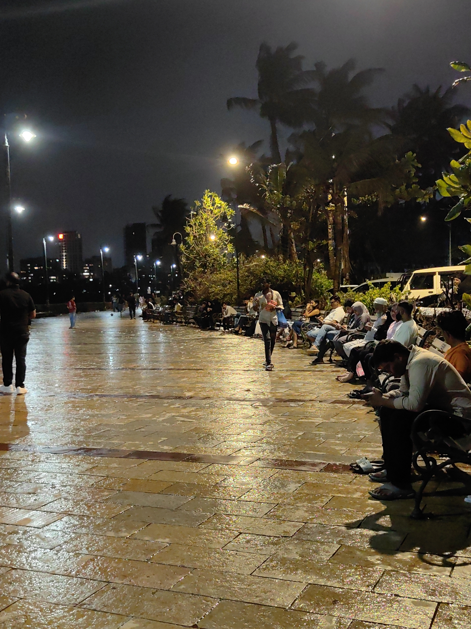 This image captures a nighttime scene at Marine Drive, a prominent promenade in Mumbai, India. The ground, paved with rectangular tiles, is wet and reflects the ambient light, suggesting recent rain. Several individuals are present, with most seated on benches along the promenade.
In the foreground, a man in a black shirt and dark pants stands facing away from the camera. Further down the promenade, a group of people are seated on benches, some looking at their phones, others engaged in conversation. A man in a white shirt walks towards the seated individuals. In the background, there are more people scattered along the promenade, with a few children playing with a ball. Palm trees and other foliage are visible on the right side of the frame, illuminated by streetlights. The skyline of Mumbai, with its tall buildings and illuminated windows, forms the backdrop. Streetlights are strategically placed along the promenade, casting a warm glow on the scene. A white van is parked in the background. The overall atmosphere is one of a calm evening by the sea.