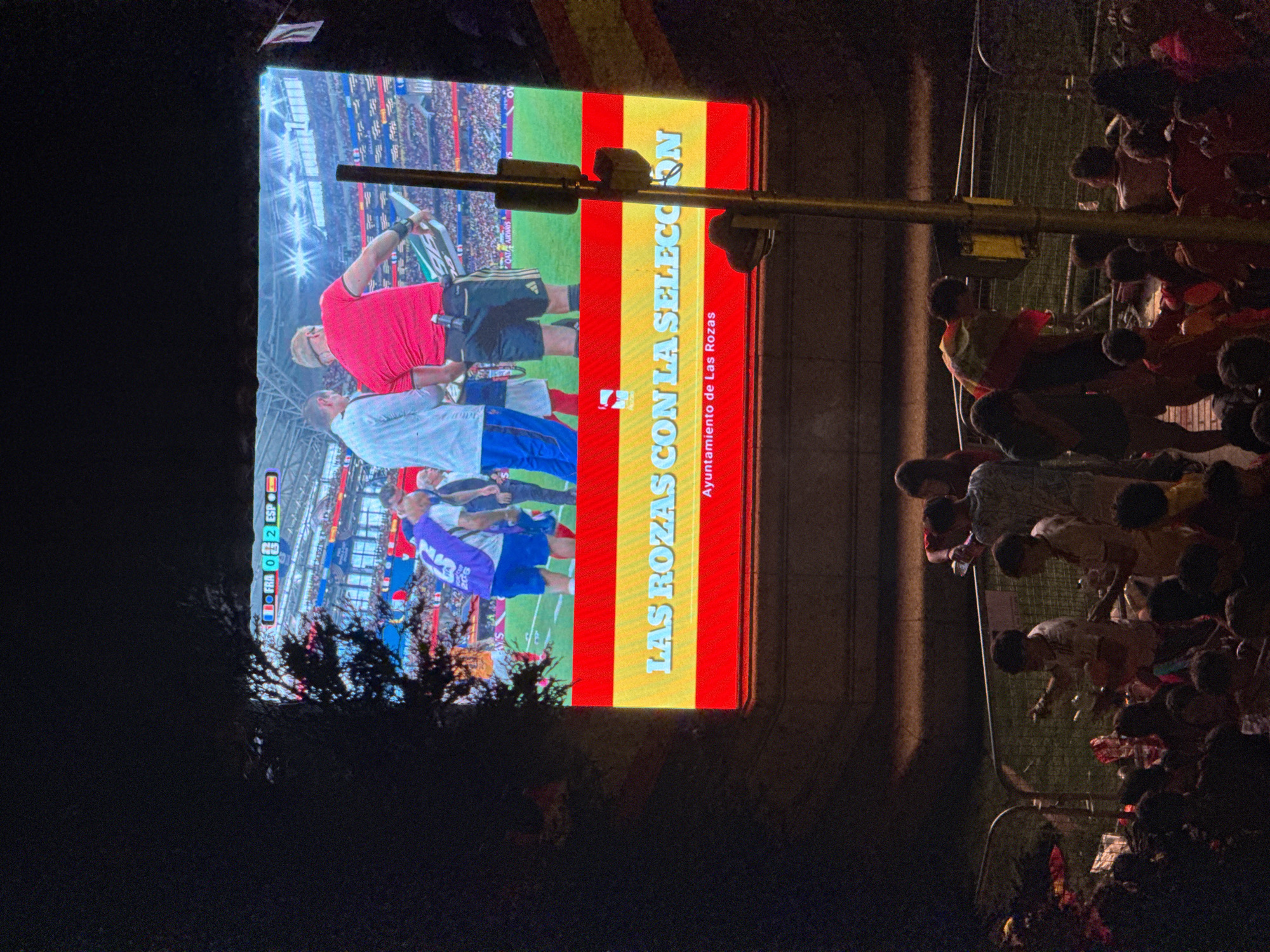 The image shows a large outdoor screen displaying a soccer game, with the score "FRA 0 2 ESP" visible. Below the game footage, a Spanish flag is displayed with the text "LAS ROZAS CON LA SELECCION" and "Ayuntamiento de Las Rozas" in yellow lettering. A crowd of people are gathered in front of the screen, some are looking at the screen, others are interacting with each other. The scene appears to be at night, with some artificial lighting illuminating the area. The presence of the "Ayuntamiento de Las Rozas" indicates the event is taking place in Las Rozas de Madrid, Spain.