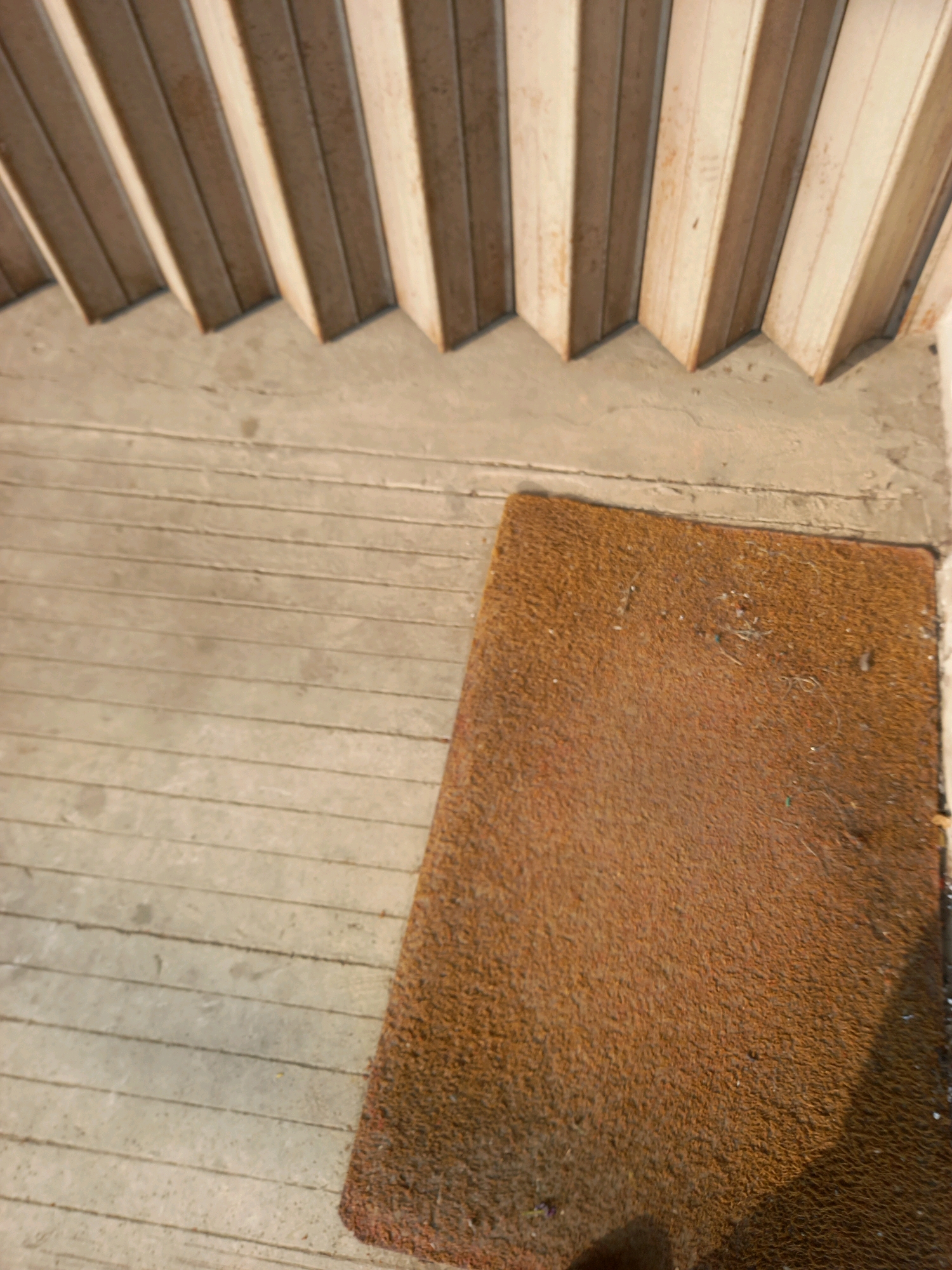 The image shows a doormat on a wooden floor. Above the floor is a set of stairs with light-colored wooden railings. The scene is outdoors, possibly in front of a building. There are no people visible in the image. The lighting suggests it is daytime. The doormat is a rectangular shape with a textured, brown surface. The wooden floor has visible planks and appears weathered. The stairs are also made of wood and have a slatted design. The overall impression is of an entranceway. There is no specific indication of its location within Samarinda, Indonesia, beyond the general context of an outdoor residential or commercial entrance.