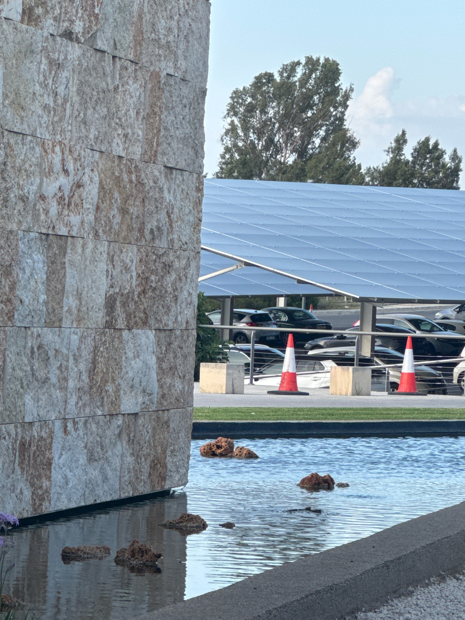 The image depicts a modern architectural element, a large, textured stone wall, situated next to a water feature with rocks. In the background, a solar panel-covered parking structure is visible, sheltering several parked cars. Two orange and white traffic cones stand in the foreground on a patch of grass, and further back, a line of trees forms the skyline against a hazy blue sky with clouds. The scene appears to be a public space, possibly a civic building or commercial complex. The location is identified as Strovolos, Cyprus.
