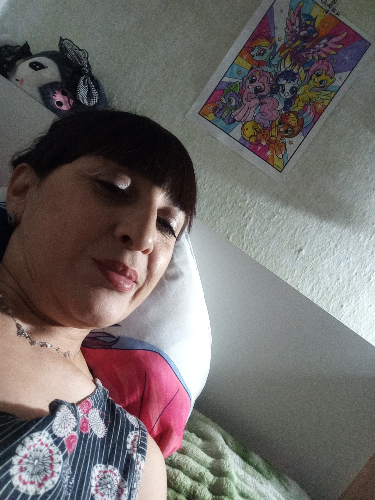 The image shows a close-up, low-angle selfie of a woman resting on a pillow with a cartoon character pillowcase. She has dark hair and bangs, and is wearing a dark blue and white striped shirt with a floral pattern and a silver necklace. To the left of the woman, a stuffed toy with a black and white bow is visible. Above her, a framed coloring page of characters from "My Little Pony" is displayed on a textured wall. The overall setting appears to be a bedroom or living space, with a green textured blanket visible in the foreground. The lighting suggests it is indoors, possibly during the day or with artificial lighting. The location of Belogorsk, Russia, is not discernible from the image itself.