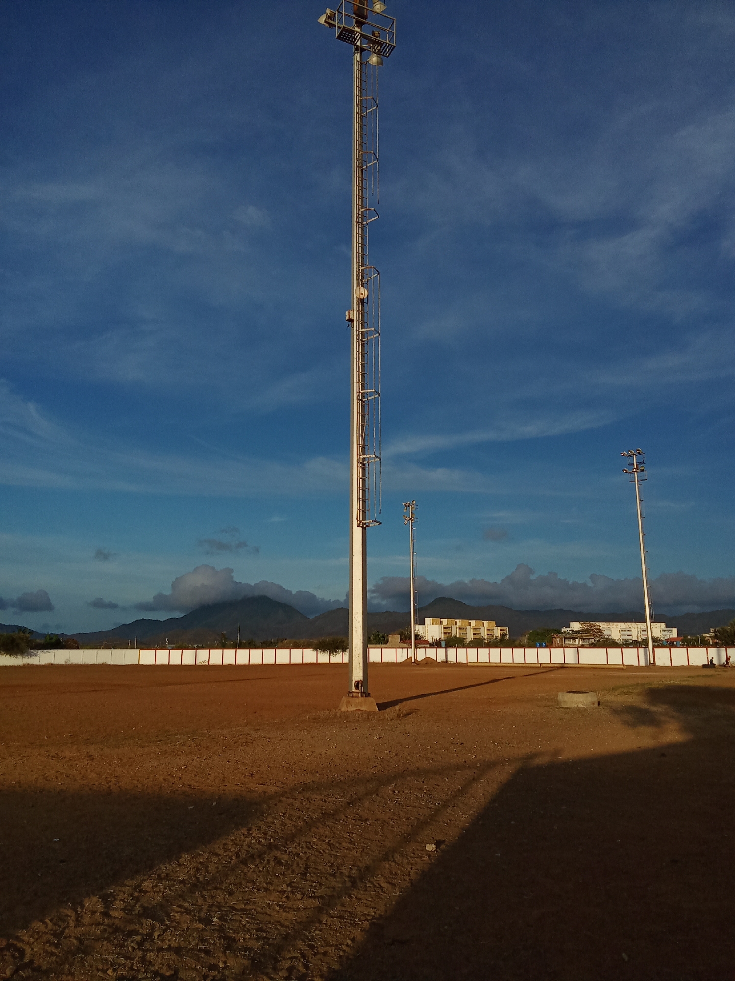 This image depicts a vast, unpaved dirt field under a bright blue sky with wispy clouds. A prominent, tall metal floodlight tower stands in the foreground, slightly to the left, casting a long, dark shadow across the reddish-brown ground towards the viewer. Two other similar, but smaller, light poles are visible further back to the right.
The field is enclosed by a low white perimeter wall with a red stripe. Beyond this wall, mountains rise in the distance, partially veiled by scattered clouds. Several multi-story buildings, appearing to be residential or commercial, are visible between the wall and the mountains.
The long shadows stretching across the field and the warm, golden light suggest the time of day is late afternoon or early evening. The sky is clear, indicating fair weather. The field appears deserted, with no specific activity taking place, though its design with floodlights suggests it could be a sports ground or a public gathering area. A small, circular concrete object rests on the ground in the middle-right. There are a few very small, indistinct human figures visible in the far distance near the rightmost light pole. No visible text is present in the image. The scene portrays a quiet, expansive landscape, possibly in Juan Griego, Venezuela.