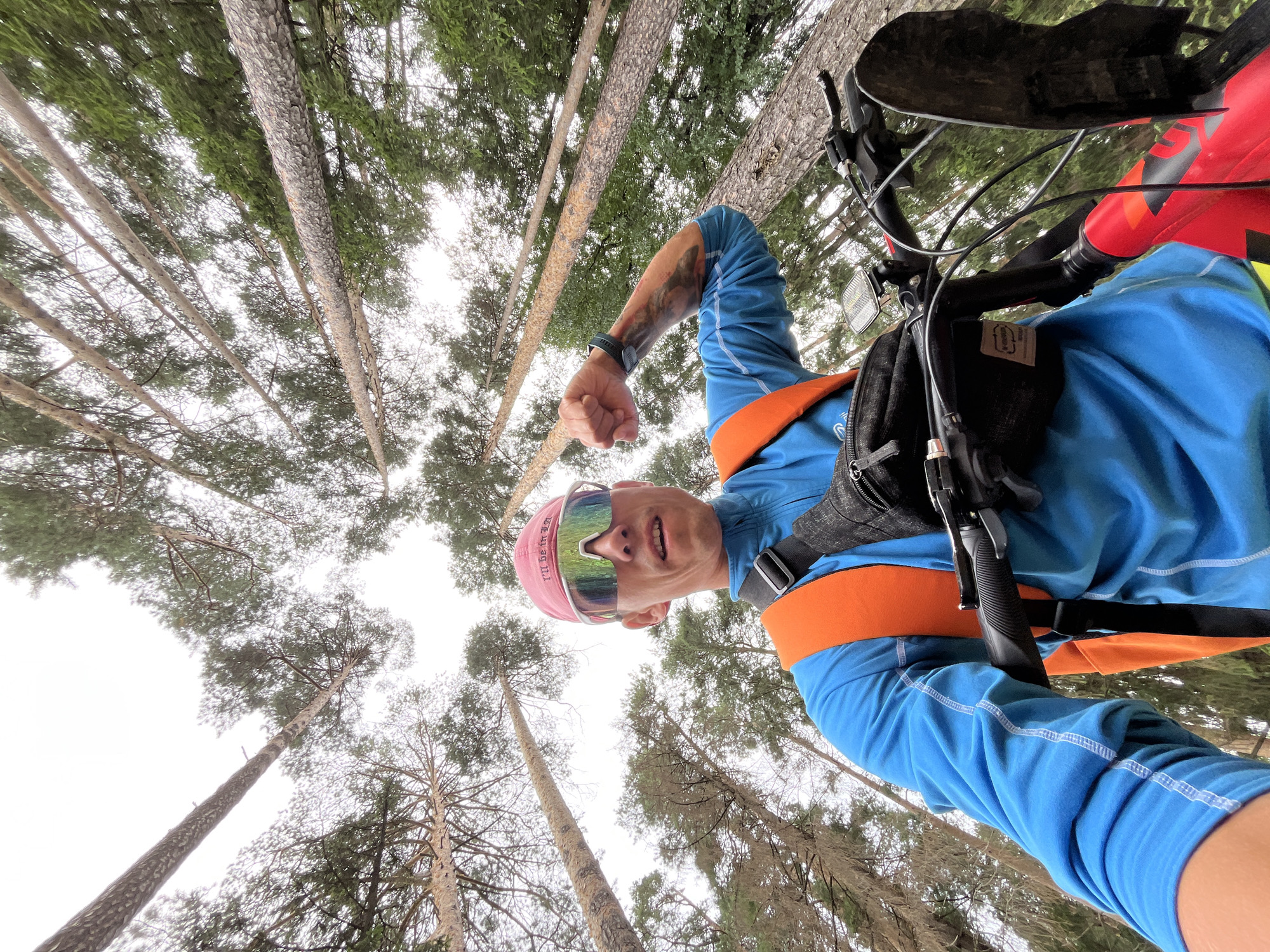 A low-angle, wide shot captures a man on a bicycle in a forest. The man, wearing a pink cap, mirrored sunglasses, a blue long-sleeved shirt, and an orange backpack, is looking at the camera with his right fist clenched. His left arm, with a tattoo, is extended. The bicycle's handlebars and part of its red frame are visible in the foreground. Tall, thin trees with green foliage dominate the background, stretching towards a bright, overcast sky. The scene is set in a forest, and based on the user's prompt, the location is specified as the city of Chaikovsky, Russia.