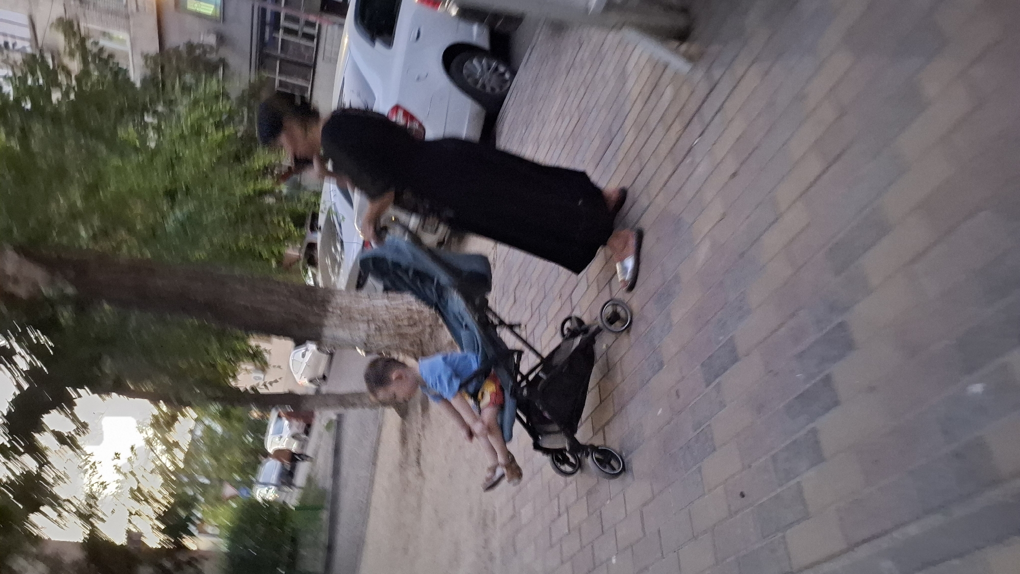 A child is in a stroller with a woman pushing it. The child is wearing a blue shirt and red and yellow shorts. The woman is wearing a black dress. They are on a paved walkway in front of a building. There are trees and cars in the background. The image is blurry and appears to be taken in Shymkent, Kazakhstan.