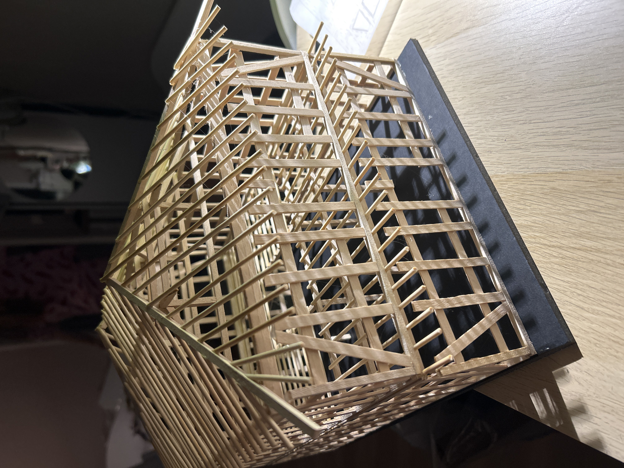 This is a close-up, angled shot of a model of a timber-framed building, likely a house or barn, constructed from thin wooden sticks. The model is built on a black base and sits on a light-colored wooden surface. The building's frame is visible, showing a two-story structure with diagonal bracing and a pitched roof frame. The model appears to be in an indoor setting, with the background out of focus and dimly lit, suggesting it might be a workbench or hobby area. There are no people or animals visible in the image, and no specific location in Sofia, Bulgaria can be determined from the visual information provided.