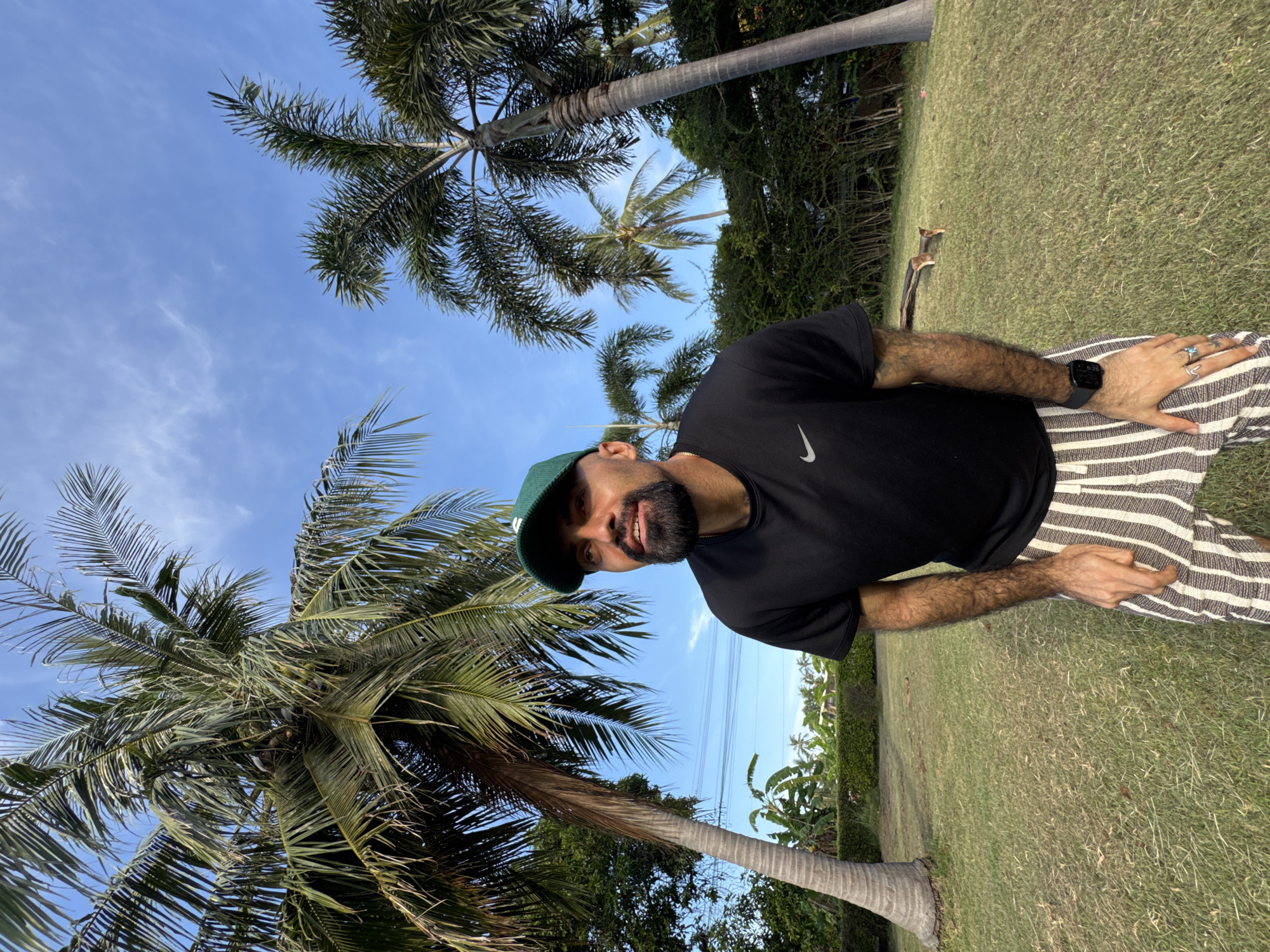 A man wearing a green baseball cap, black t-shirt with a Nike logo, and striped shorts stands in a grassy outdoor area with palm trees and lush greenery in the background. The sky is blue with scattered clouds. The man is looking up and to his right, with his hands resting on his upper thighs. The image appears to have been taken in the Ko Samui District of Thailand.