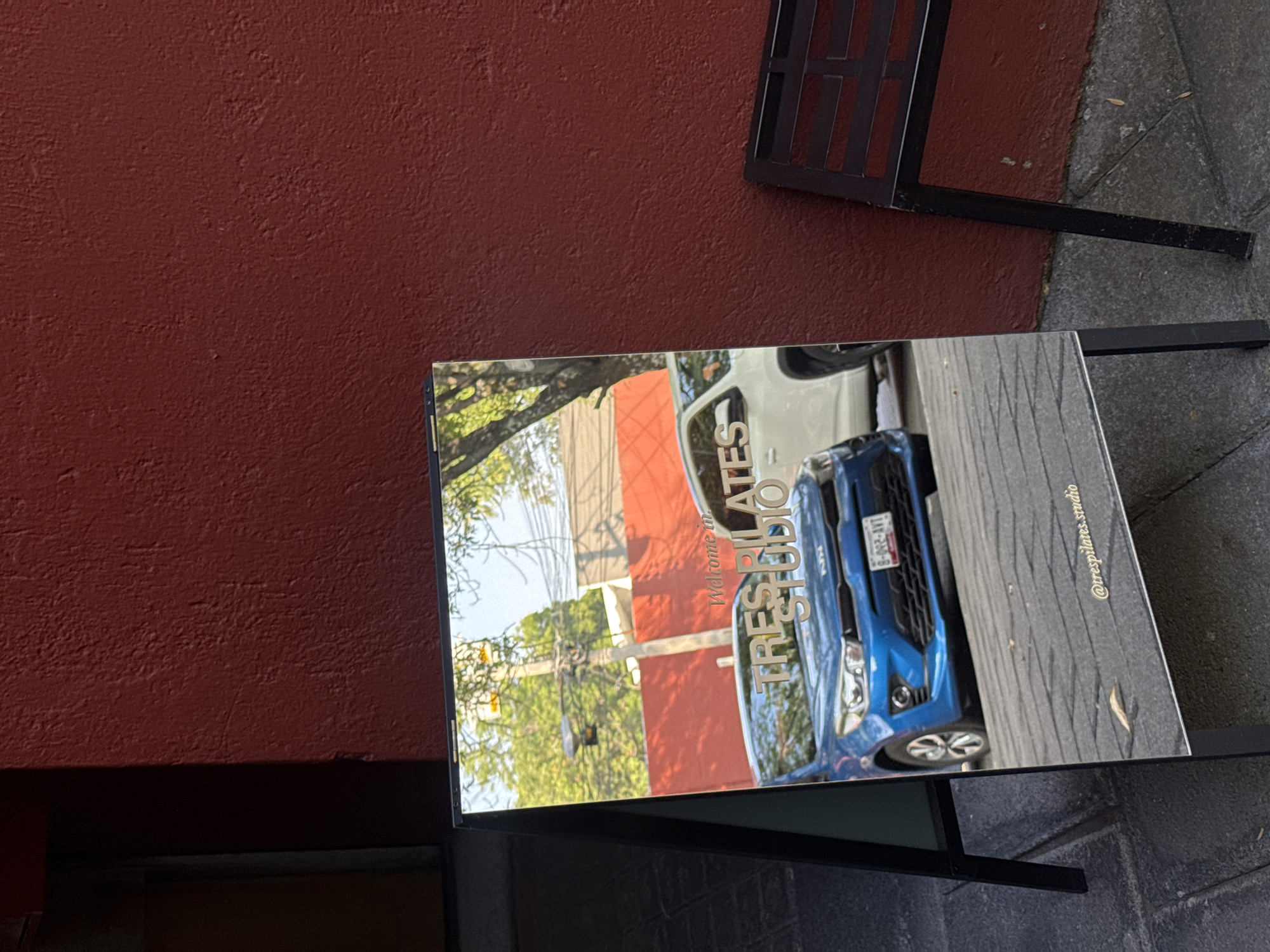 The image shows a metal advertising easel with an image of two cars, one blue and one white. The image reads "Welcome to TRES PILATES STUDIO" with the social media handle @trespilates.studio in the lower right corner. The easel stands on a grey sidewalk next to a dark red wall. Part of a metal chair is in the upper right corner. The scene is in Puebla, Mexico.