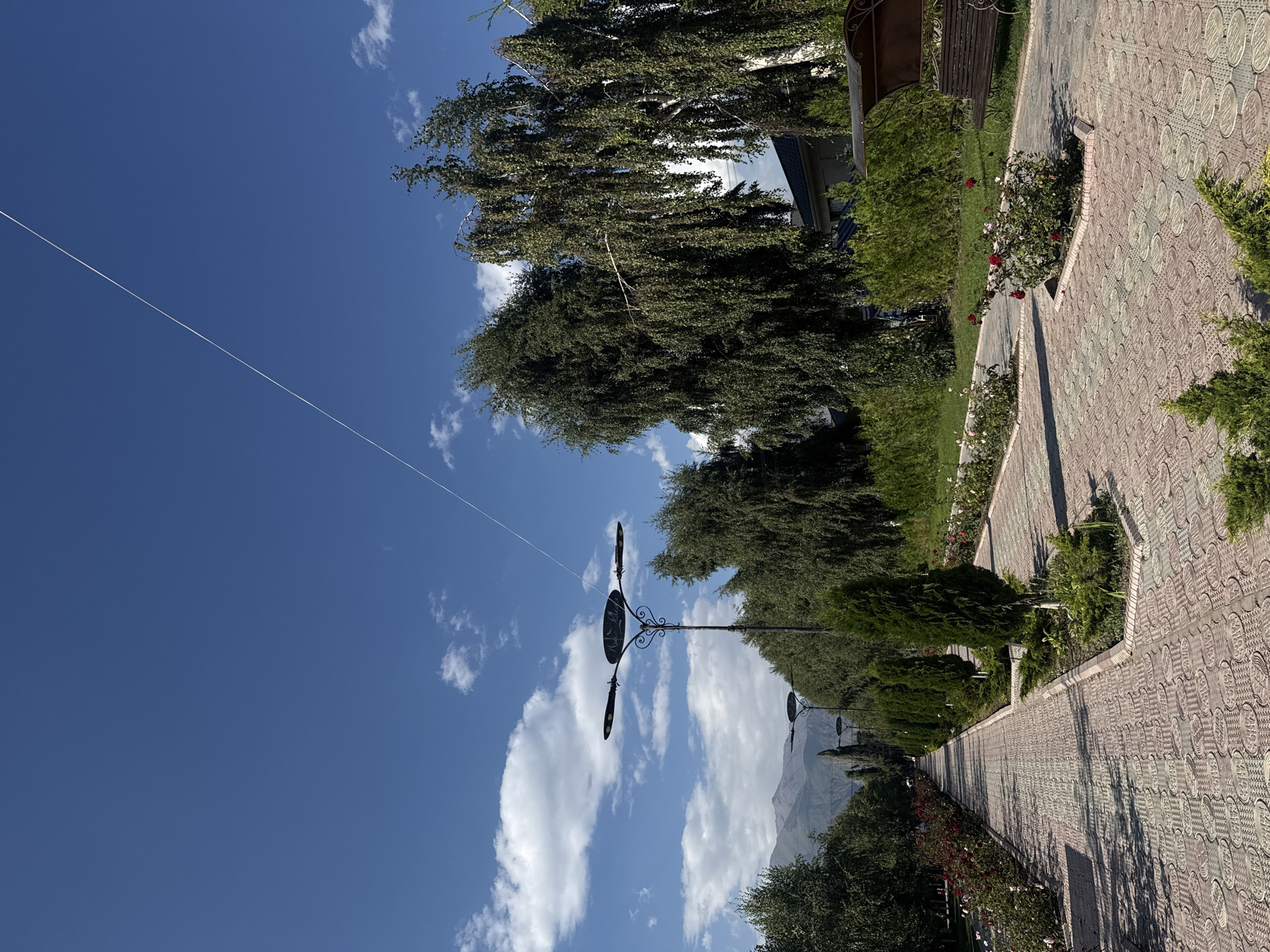 A long, patterned brick pathway leads into the distance in a park-like setting. The pathway is lined with rose bushes and manicured evergreen trees. In the background, tall trees with drooping branches, possibly birch, are visible, along with a building. A thin, white line stretches diagonally across the bright blue sky, which is dotted with white clouds. Mountains are visible in the far distance. A decorative lamppost stands alongside the path, and a bench with a canopy is visible on the right side of the image. The scene appears to be a public outdoor space in Sary-Oy, Kyrgyzstan, likely a park or pedestrian area designed for recreation.