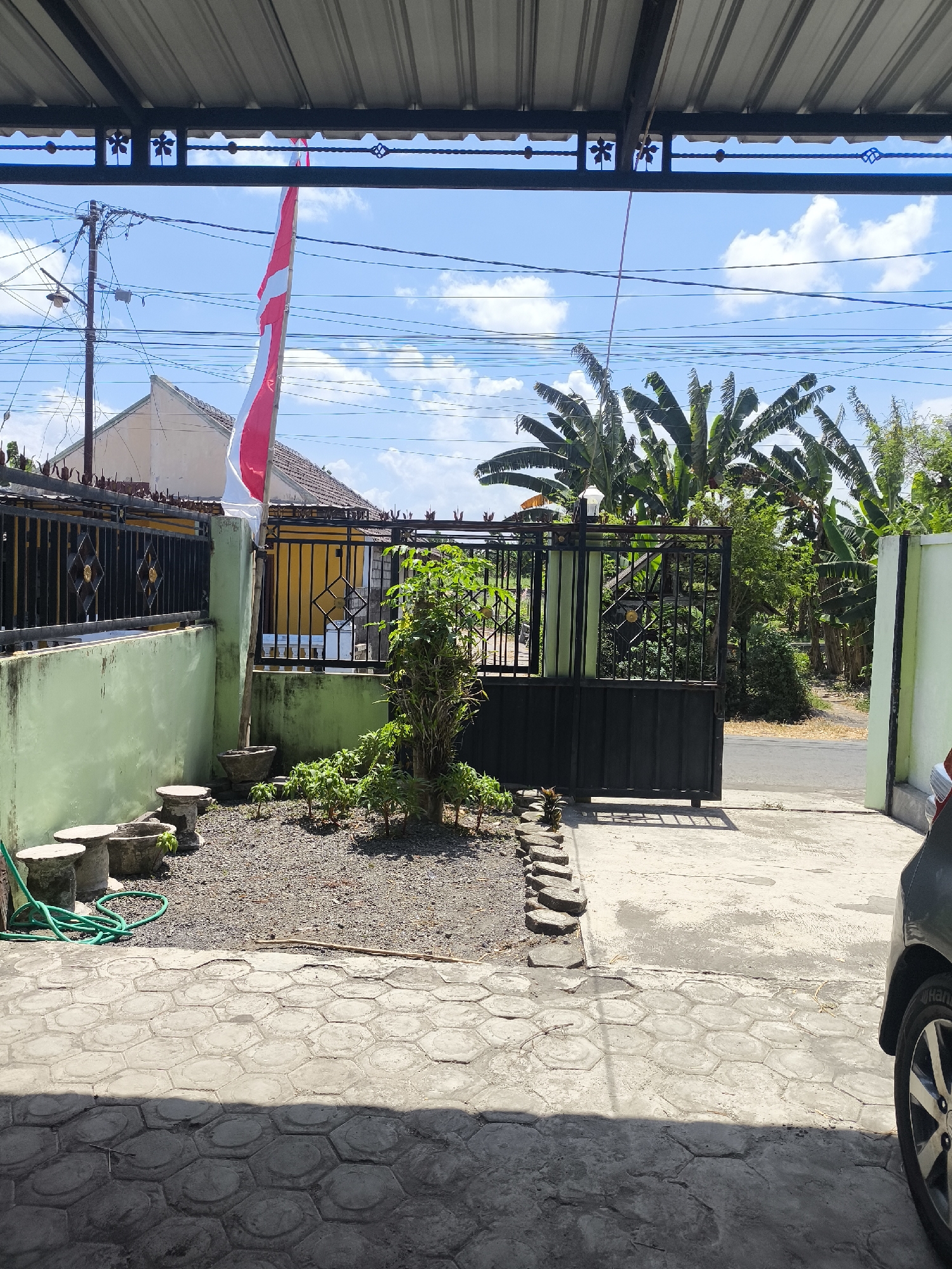 The image depicts a residential outdoor scene in Kebonsari, Indonesia, on a clear day with scattered clouds. In the foreground, a grey car is partially visible on the right side, with its rear wheel and fender in view. The ground is paved with hexagonal concrete tiles. To the left, a small, gravel-covered yard features several concrete planters and a coiled green garden hose. A young, leafy plant stands in the center of the yard, near a black metal gate. Behind the gate, banana trees and other greenery are visible. A red and white Indonesian flag is prominently displayed on a pole to the left of the gate, attached to a light green wall. A black wrought-iron fence runs along the wall. The background shows houses with tiled roofs, power lines, and a utility pole. The scene appears to be a view from a covered porch or carport, indicated by the corrugated metal roof structure overhead.