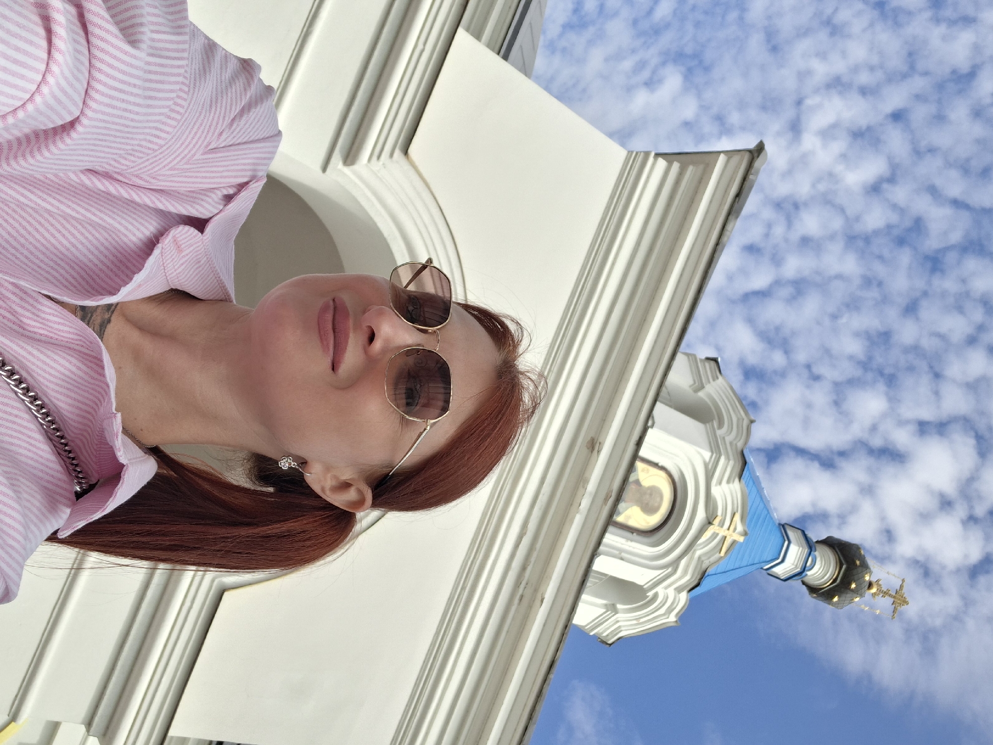 A woman with auburn hair tied in a ponytail, wearing a pink and white striped shirt and sunglasses, takes a selfie in front of a white building with a blue-domed roof and a golden cross. The building appears to be a church or chapel, possibly located in the city of Pastavy, Belarus. The sky is bright blue with scattered white clouds. The woman is smiling slightly and looking towards the camera. She is wearing a small silver earring in her left ear. A silver chain strap from a bag is visible across her shoulder. The perspective is low-angle, emphasizing the architecture of the building and the sky.