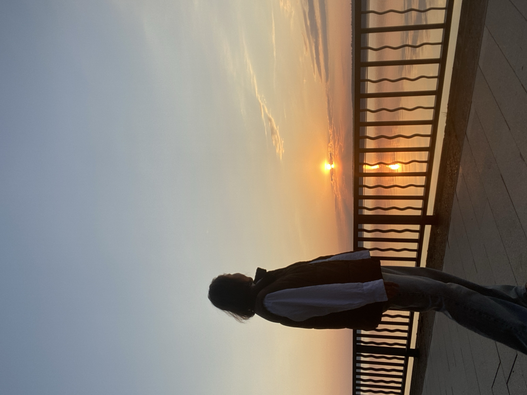 A woman is standing next to a metal railing, silhouetted against a sunset. The sky transitions from light blue to orange near the horizon. The sun is reflected in the water behind the railing. The woman is wearing a jacket and jeans. The railing is on a paved surface. The scene appears to be near water with a sunset. The location is likely in the city of Chaikovsky, Russia.