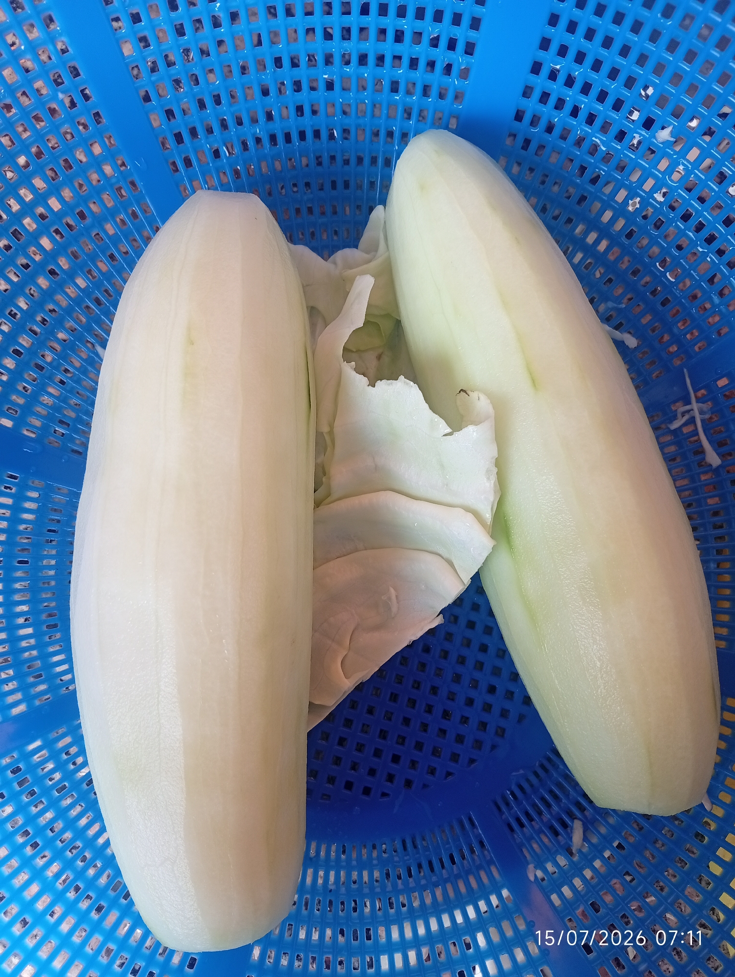 Two peeled white cucumbers rest on a blue plastic basket. A few shredded pieces of green cabbage are also visible in the basket, tucked between the cucumbers. The basket's grid pattern is clear, and some water droplets are visible on its surface. The timestamp "15/07/2026 07:11" is overlaid on the bottom right corner of the image. The location is the city of Bana Stream, Nigeria.