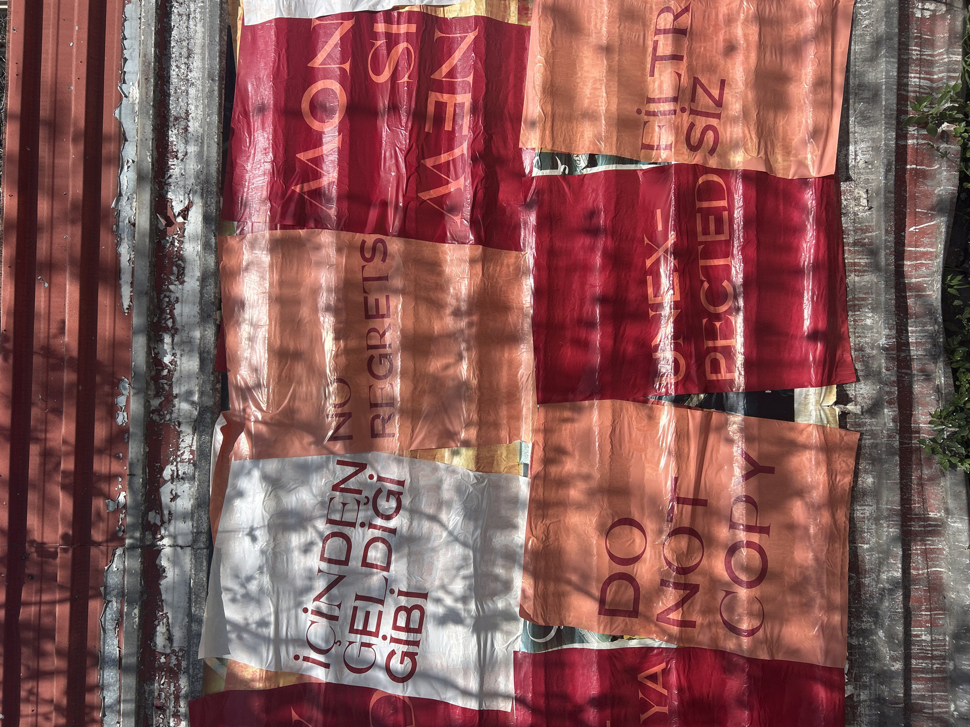 The image presents a collection of plastic bags laid out on what appears to be a corrugated metal surface. The surface shows signs of weathering and peeling paint, suggesting it is an outdoor structure or roof.
The plastic bags feature various printed messages in different colors, primarily red and orange on a white or light-colored background. The messages include English phrases like "NEW IS NOW," "NO REGRETS," and "DO NOT COPY." One bag features the Turkish phrase "İÇİNDEN GEL DİĞİ GİBİ," which translates to "As it comes from within." Another says "FILTER SIZ EX-PECTED"
The environment seems to be outdoor, as suggested by the presence of plant life in the background on the right side. Shadows cast across the scene suggest the presence of sunlight.
While the image alone doesn't definitively locate the scene in Beyoğlu, Turkey, the presence of the Turkish phrase on one of the bags suggests a Turkish context, so location is reasonable.