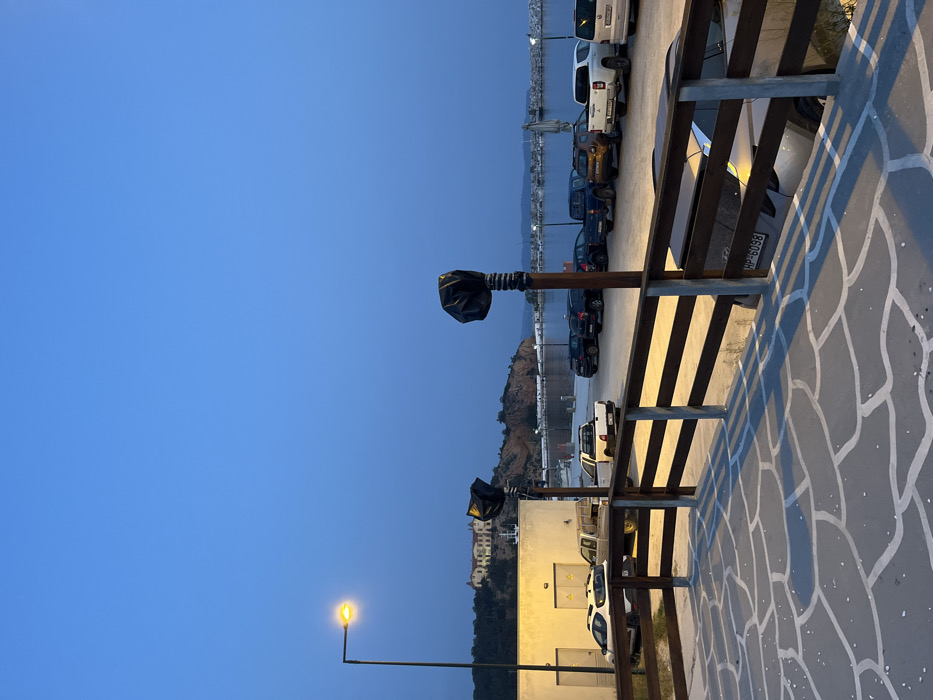 The image shows a view from a patio in Limenaria, Greece (latitude: 40.626, longitude: 24.575). The patio is made of gray stone tiles with white lines, and it is surrounded by a wooden railing. The patio faces a parking lot with a row of parked cars and trucks. There is a small building on the left, a hill in the background, and a body of water in the distance. The sky is clear blue and the sun is setting. There is a street light on the left and two more lights in the foreground. There are no people in the image.