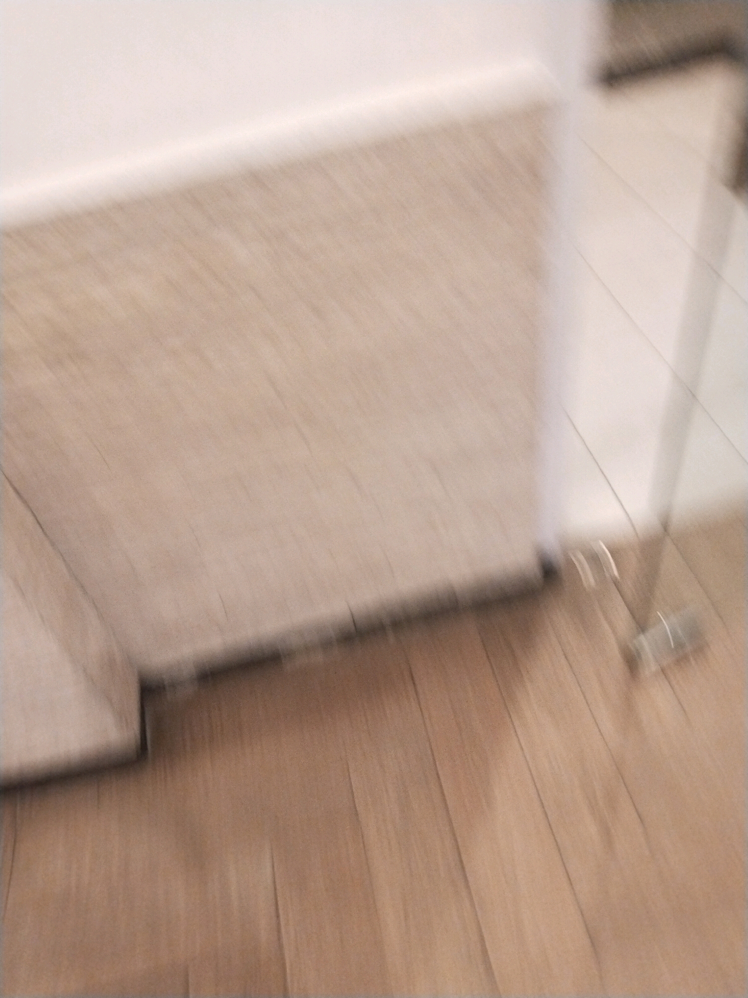 The image depicts a floor with wooden planks and a baseboard. In the lower right portion of the image, there are two transparent cylindrical objects that appear to be bottles. One bottle is upright, while the other is lying on its side. The lighting suggests an indoor setting. The image is blurred due to motion, making it difficult to discern finer details or the specific location within Surakarta, Indonesia.