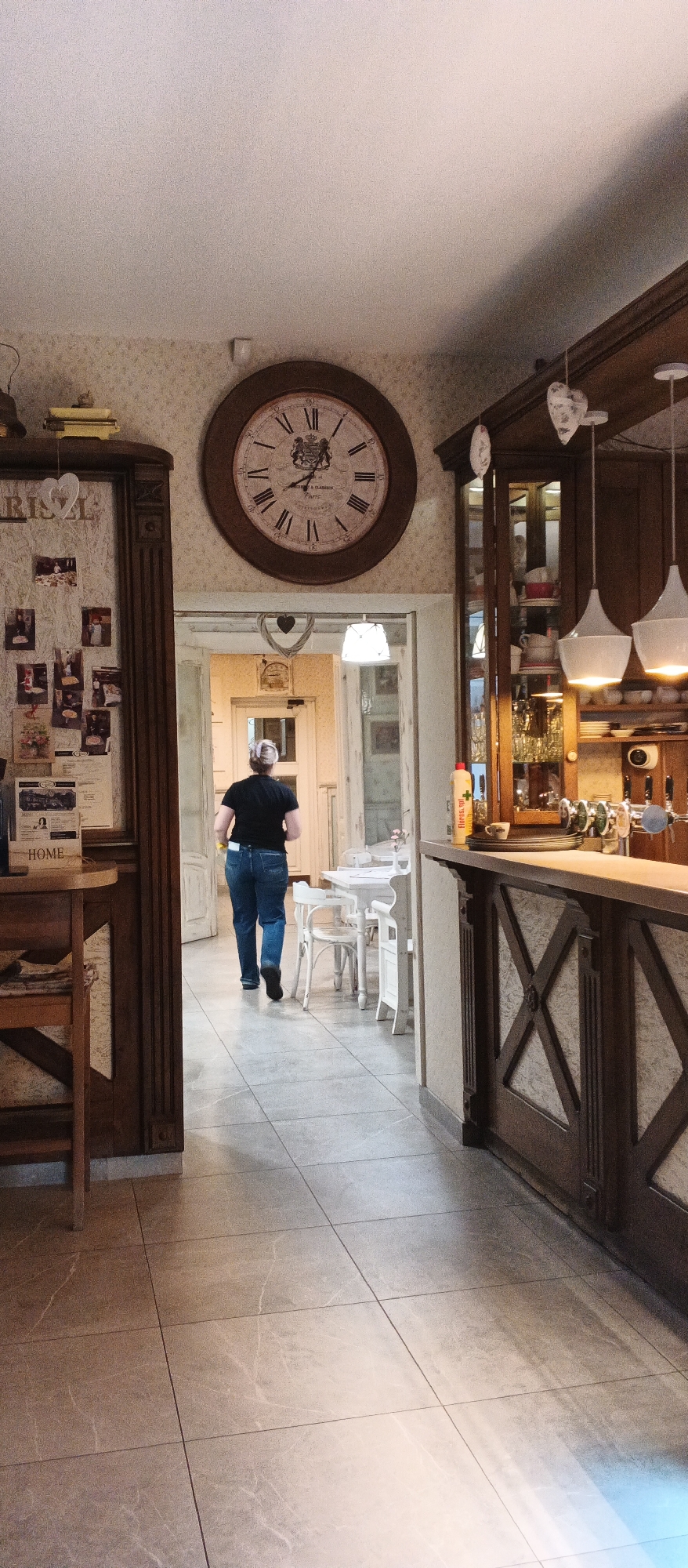 A person wearing a black t-shirt and blue jeans walks away from the camera into a brightly lit hallway. The person's blonde hair is pulled up in a bun. They are walking on a tiled floor that appears to be in a restaurant or cafe. To the left of the hallway is a large wooden cabinet with pictures and a clock on the wall above it. To the right of the hallway is a bar area with a wooden counter and shelves stocked with various items. The lighting is warm and the overall atmosphere appears to be cozy and inviting. The restaurant is likely located in Berehove, Ukraine, a city known for its thermal spas and wineries.