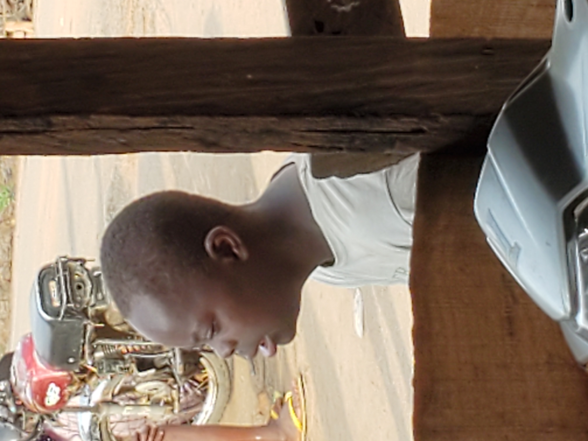 An eye-level shot captures a young individual with short hair wearing a white shirt. The person appears to be looking down. A motorcycle is partially visible to the left, and a white scooter is to the right. The foreground is framed by a wooden structure, possibly a porch or awning. The scene is likely located in Mararaba, Nigeria, given the request.
