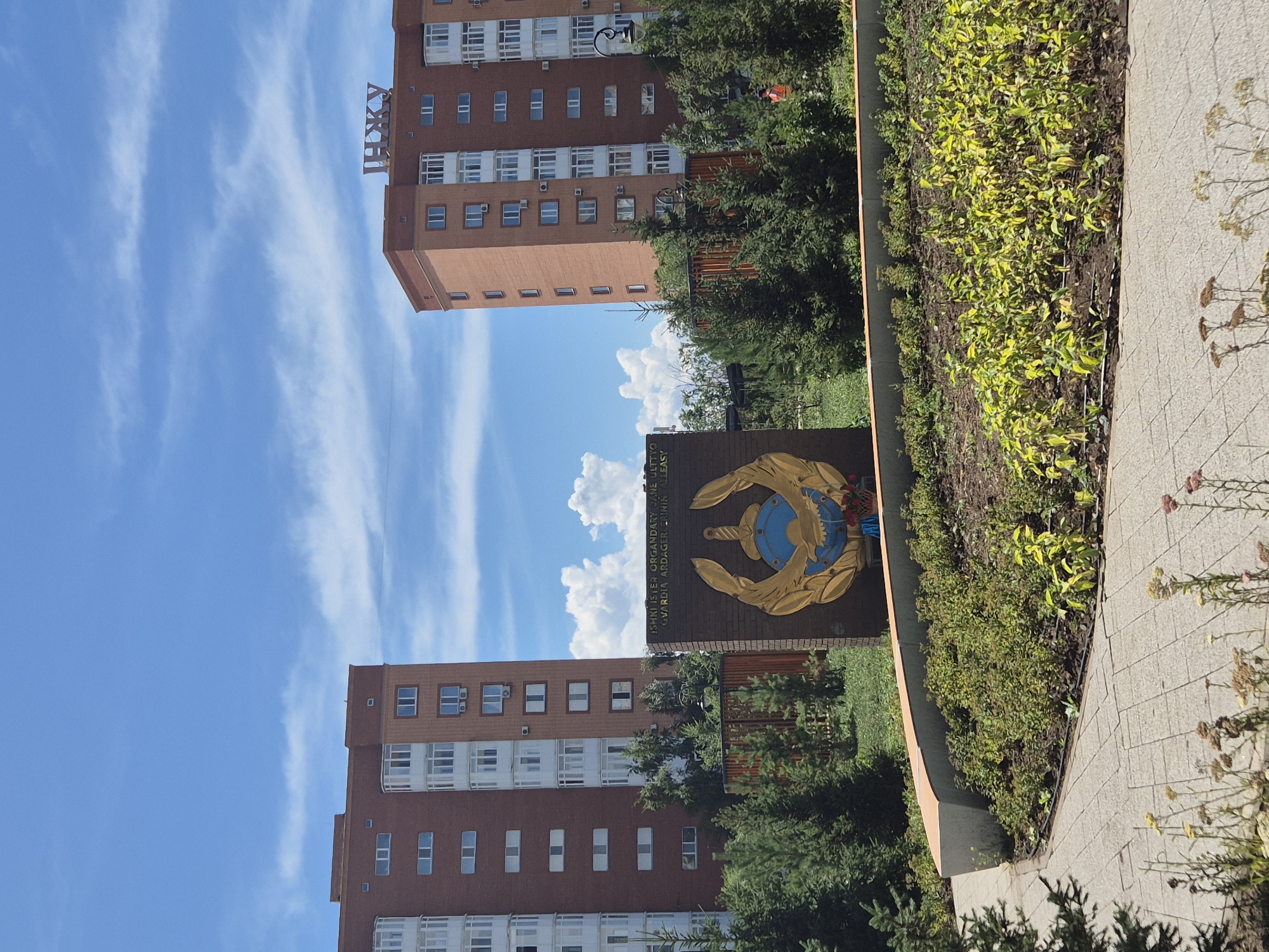 The image shows two tall, reddish-brown apartment buildings under a bright blue sky with scattered white clouds. The building on the left is larger and appears to be an older style, while the one on the right is slightly smaller and has a more modern appearance, featuring a sign with Cyrillic lettering that reads "ИНЖУ" on its roof. In the foreground, there is a monument or memorial made of dark brick, adorned with a gold emblem featuring a sword, a globe, and laurel leaves. The text on the memorial is partially visible and appears to be in a language using Latin script, possibly related to a military or governmental organization. The area around the memorial is landscaped with green bushes and trees, and a paved walkway made of square grey tiles is visible in the lower part of the frame. The overall scene depicts a public space in a city, likely a residential area given the apartment buildings. The location is identified as Kokshetau, Kazakhstan.