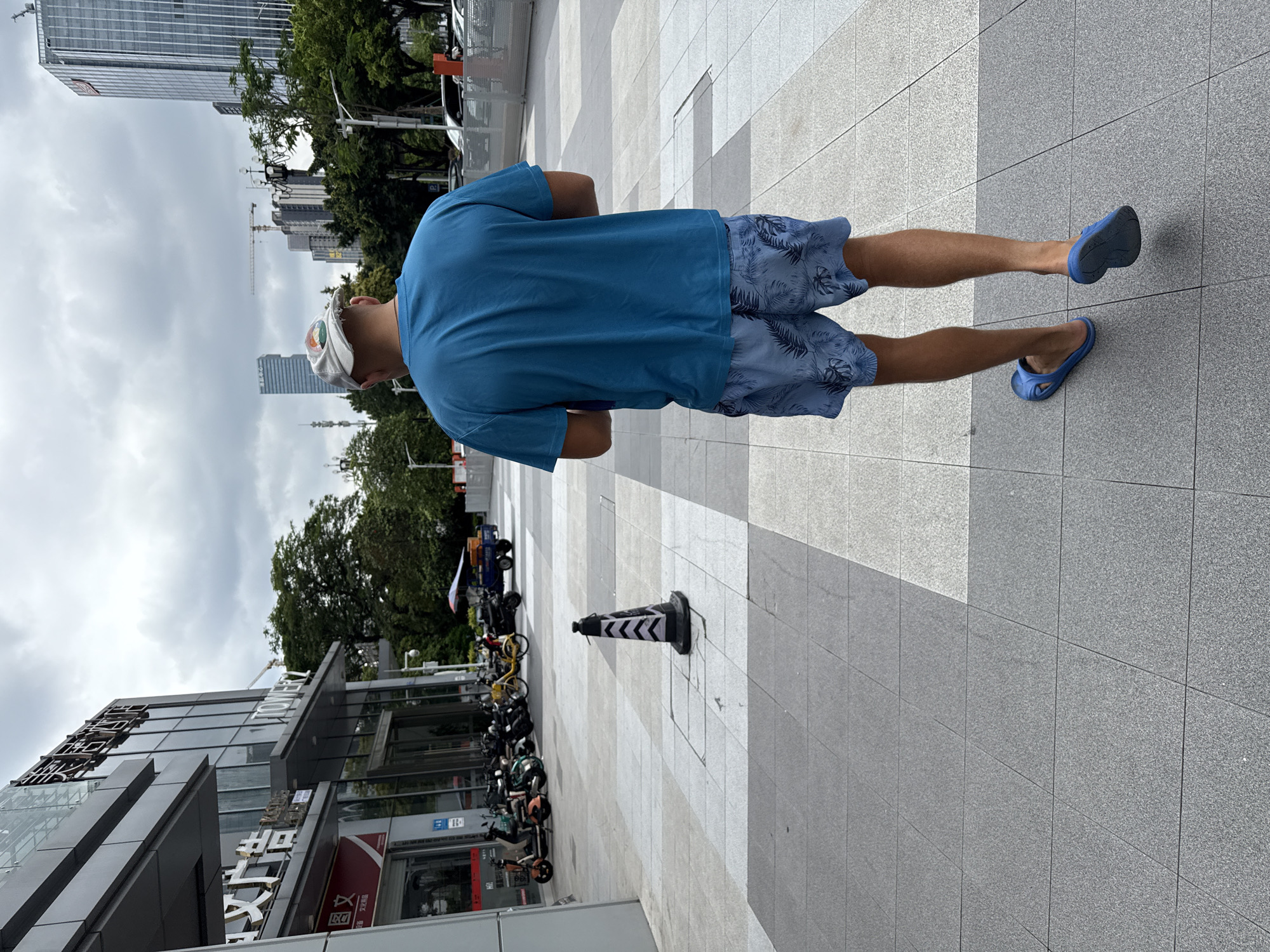 In Huangpu District, China, a man wearing a blue t-shirt and blue patterned shorts walks away from the camera on a paved walkway. He is also wearing blue flip-flops. In the background, a modern building with glass windows and signage is visible. To the left, several electric scooters are parked. Further in the distance, tall buildings and construction cranes indicate an urban setting. The sky is overcast. A black and white traffic cone is positioned on the walkway in the foreground.