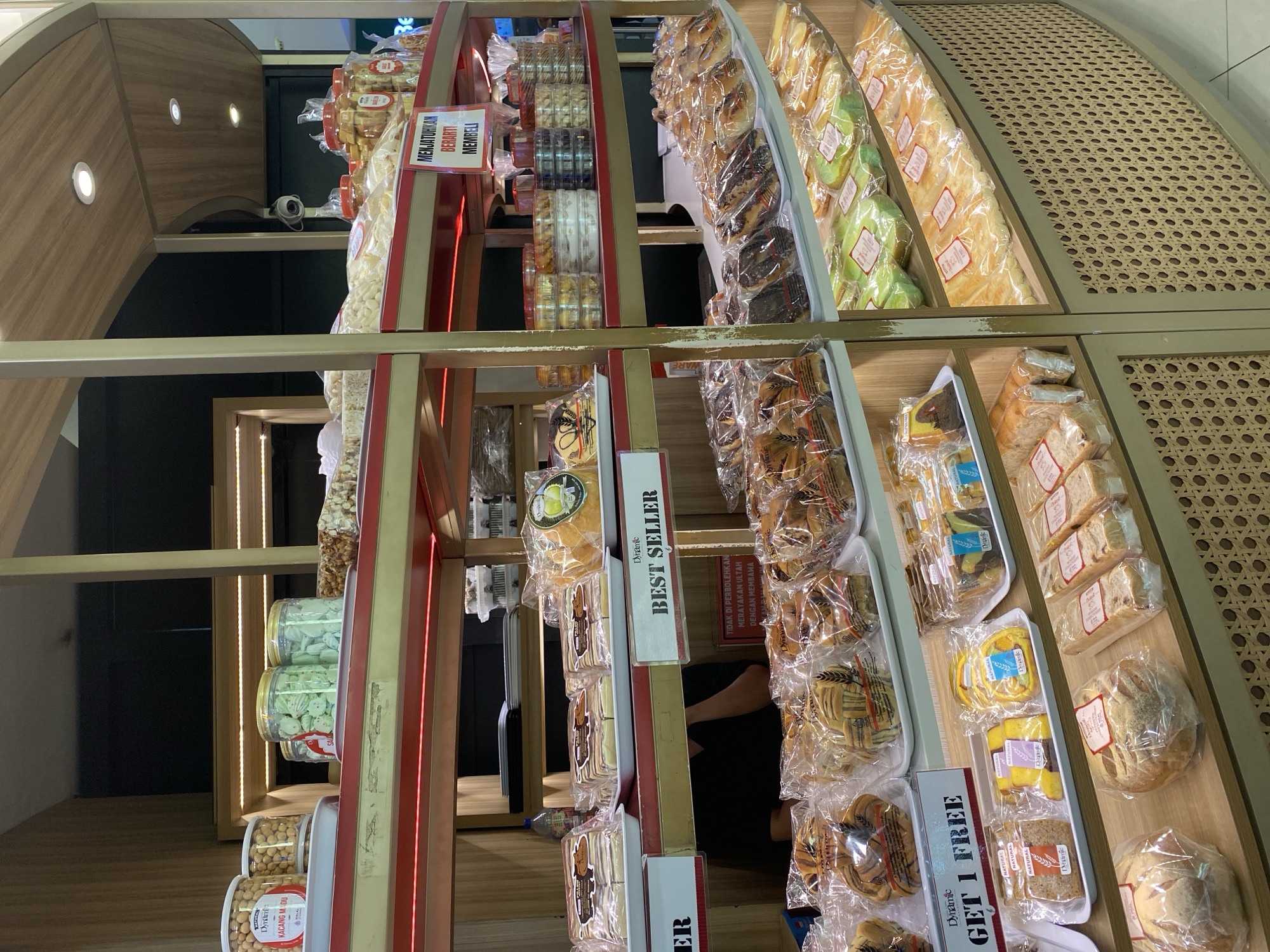 This is a photograph of a bakery display, likely taken in West Jakarta, Indonesia, given the Indonesian text on the signage. The bakery features multiple shelves laden with various baked goods and packaged snacks.
On the top shelf, several large plastic containers filled with what appear to be cookies or crackers are visible. One container, labeled "Dinamik Kacang Madu," contains honey peanuts. To the right, there are more containers of snacks, some with gold-colored lids.
The middle shelves display a variety of packaged baked goods. On the left, rows of individually wrapped cakes or pastries are arranged on white trays. A sign in the center reads "Dynamic BEST SELLER." To the right of this, more packaged baked goods are displayed, including some with prominent branding like "Dynamic."
The lower shelves showcase more baked items. On the left, there are packaged breads and pastries, with one prominent label indicating "GET 1 FREE." Further to the right, the shelves are filled with an assortment of cakes and breads, all individually wrapped in plastic. Some of these appear to be loaf cakes or layered pastries.
The overall environment suggests a retail setting, likely a supermarket or a dedicated bakery outlet. The display is well-lit, and the products are neatly arranged. The signage in Indonesian indicates the language of commerce and is common in retail establishments in Indonesia.