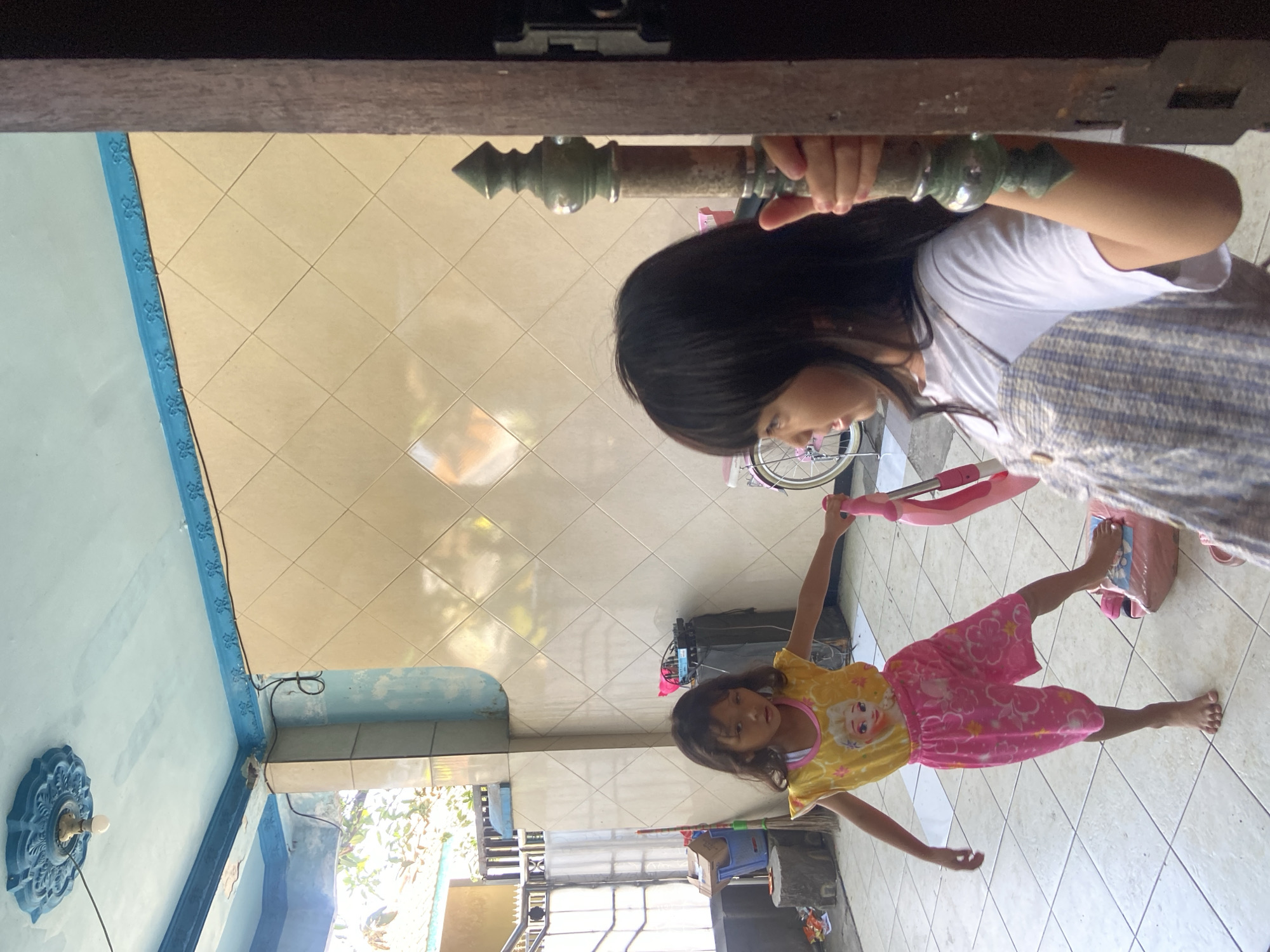 The image depicts two young girls on the tiled floor of an outdoor or semi-outdoor area in Gelam, Indonesia. The girl on the left, wearing a yellow t-shirt with an image of Elsa from Frozen and pink patterned shorts, is standing with her right foot on a pink toy, possibly a scooter or ride-on toy. Her left arm is extended towards the girl on the right. The girl on the right, wearing a white t-shirt and a blue and white striped pinafore, is partially obscured by a wooden door frame. She is holding onto a dark green, ornate metal pole that appears to be part of the door or a decorative element. In the background, a light blue ceiling with decorative molding is visible, along with cream-colored tiled walls. There are also glimpses of outdoor elements like foliage and a fence. The overall impression is of a candid moment, possibly during playtime.