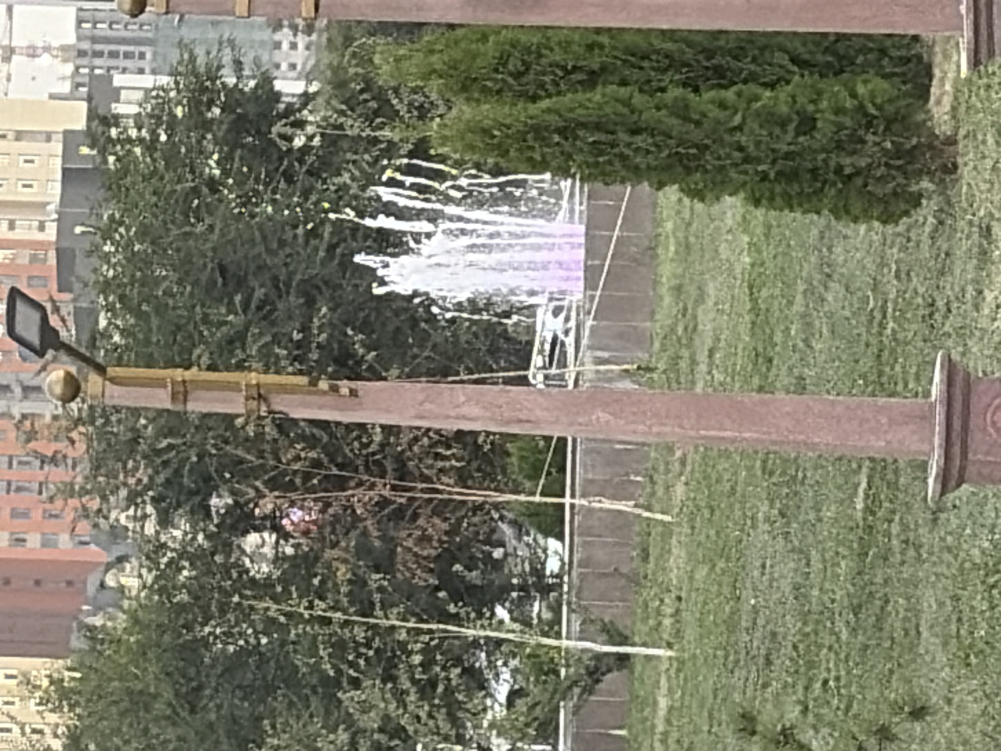 A public park scene features a prominent, reddish-brown lamppost in the foreground, casting a shadow on the green grass. Behind it, a fountain sprays water, illuminated with purple light. Tall trees and manicured bushes are scattered throughout the park. In the background, apartment buildings of varying heights and architectural styles can be seen, with a construction crane also visible. The sky is overcast, suggesting a cloudy day.