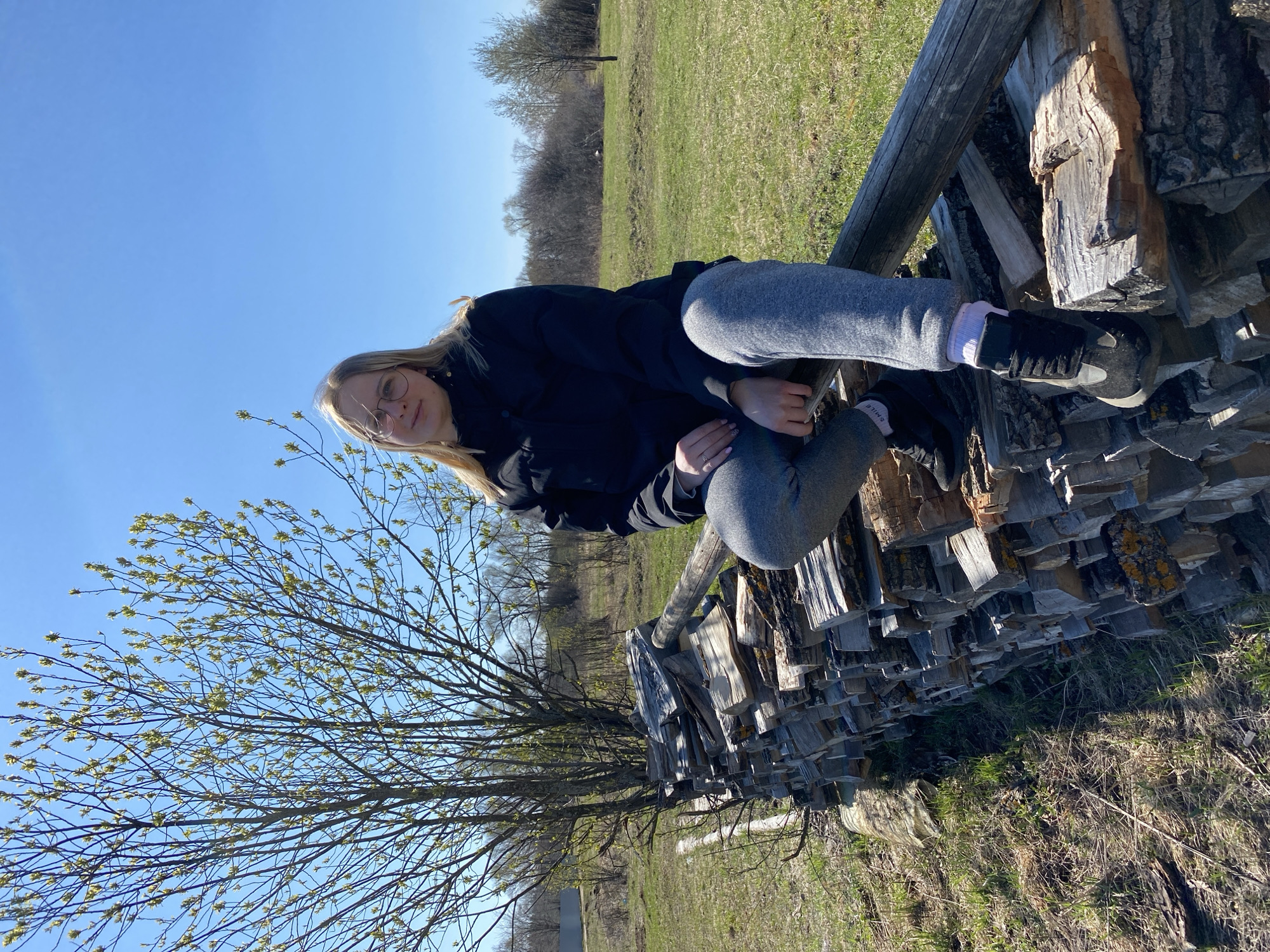 A young woman wearing glasses, a black jacket, and gray sweatpants is sitting on a stack of firewood in a grassy field. The field is partially green and partially brown. A tree with budding leaves is to the left of the firewood. The sky is blue. The location is likely outdoors, possibly in a rural area. The city of Kauchuk Stantsiya, Russia, cannot be determined from the image.