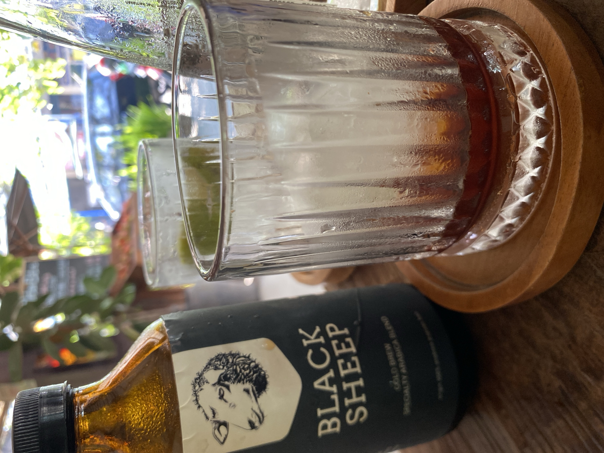 A close-up, eye-level, outdoor shot features a bottle of "BLACK SHEEP COLD BREW SPECIALTY ARABICA BLEND" and two glasses of drinks on a wooden table. The bottle, with a black cap and label depicting a sheep's head and text, is on the left. To its right, a glass with ice and a dark liquid is in the foreground, and another glass with ice and a lighter, greenish liquid is partially visible behind it. The background is blurred, showing foliage, a car, and buildings, suggesting an urban setting in North Kuta, Indonesia. The lighting indicates it is daytime, possibly morning or afternoon, with sunlight filtering through.