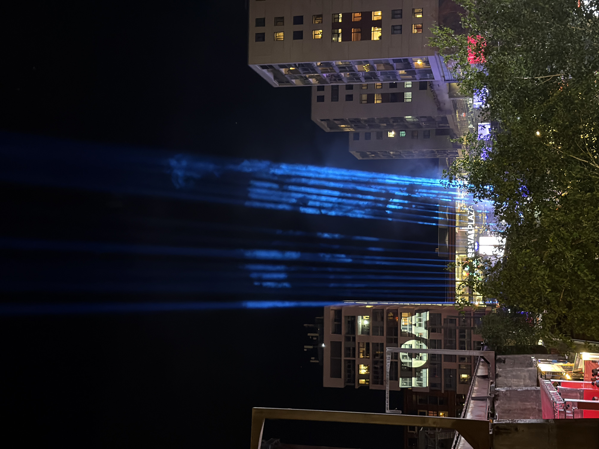 The image depicts a night scene in Esch-sur-Alzette, Luxembourg, with a view of Belval Plaza. Multiple buildings with illuminated windows are visible, some of which are partially obscured by tree foliage. Light beams emanate from the area of Belval Plaza, projecting horizontally across the dark sky. A foreground includes metallic structures and hints of red surfaces, suggesting elements of urban architecture or temporary installations.