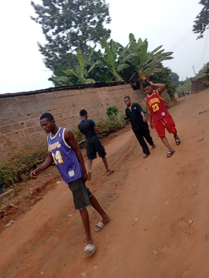 Four young men stand on a dirt road near a brick wall. One wears a Lakers jersey and cut-off shorts with sandals, another a black shirt and shorts. A third man in a black suit holds a phone, and the fourth wears a Cleveland Cavaliers jersey and red shorts. Banana trees and other foliage grow nearby. The sky is overcast.