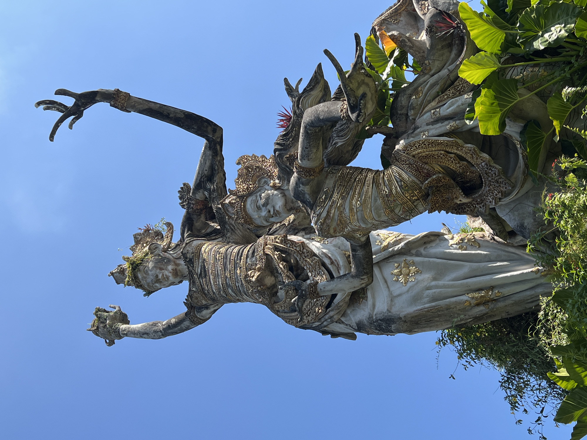 The image shows a stone sculpture in Kedewatan, Indonesia. Two figures are entwined; the top figure is smaller and appears to be riding on the larger figure's back. Both figures are adorned with elaborate headdresses and decorative garments. The sculpture exhibits some weathering and is partially covered in moss and foliage. The background is a clear blue sky. To the right, green leaves are partially covering the sculpture.