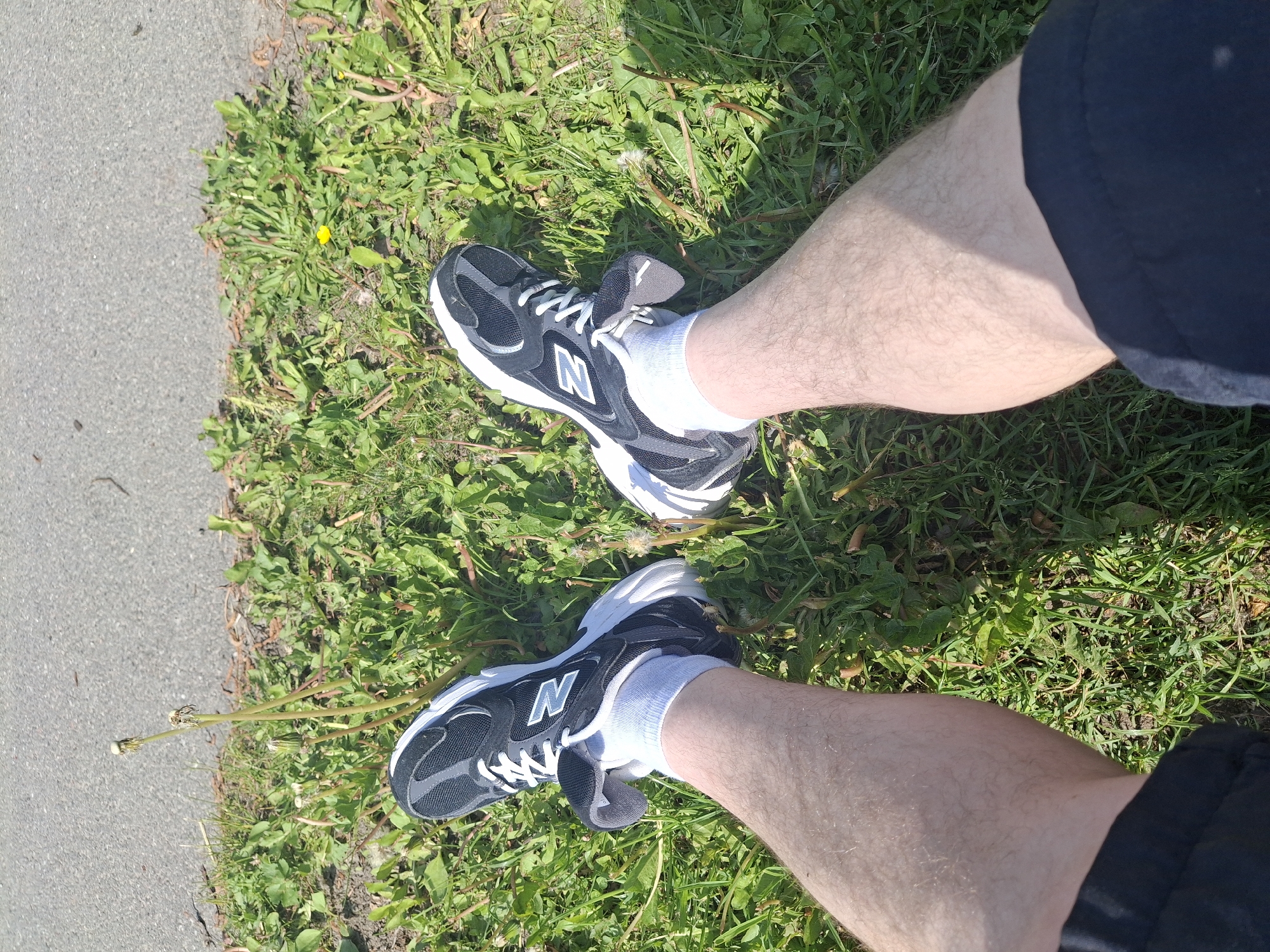 This media file captures a top-down view of a person's legs and feet resting on a grassy verge next to an asphalt path in Sillamäe linn, Estonia. The subject's bare legs are light-skinned and partially visible, extending away from the viewer. The person is wearing black and white New Balance sneakers, each featuring a distinct white "N" logo on the side, paired with white ankle socks.
The setting is outdoors on a bright, sunny day, evidenced by the strong sunlight and visible shadows cast on the grass and subtly on the legs. The ground consists of a textured grey asphalt path on the left, bordering a lush green area of grass and various weeds, including dandelions, some with small yellow blossoms and others in their white, seeding stage. The overall scene suggests a moment of rest or pause in an open, natural-urban environment, possibly a park or a pathway. No specific activity or emotion is discernible beyond the static pose of the legs and feet.