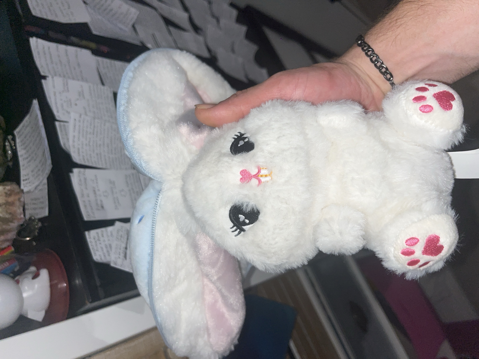 In an indoor environment, a person's hand holds a fluffy white plush bunny. The bunny has large, floppy light blue and pink ears, and embroidered facial features including black eyes with eyelashes, and a small pink nose and mouth. The bunny's paws have pink paw prints. The background is cluttered with papers and other objects on shelves. The hand is wearing a silver chain bracelet.