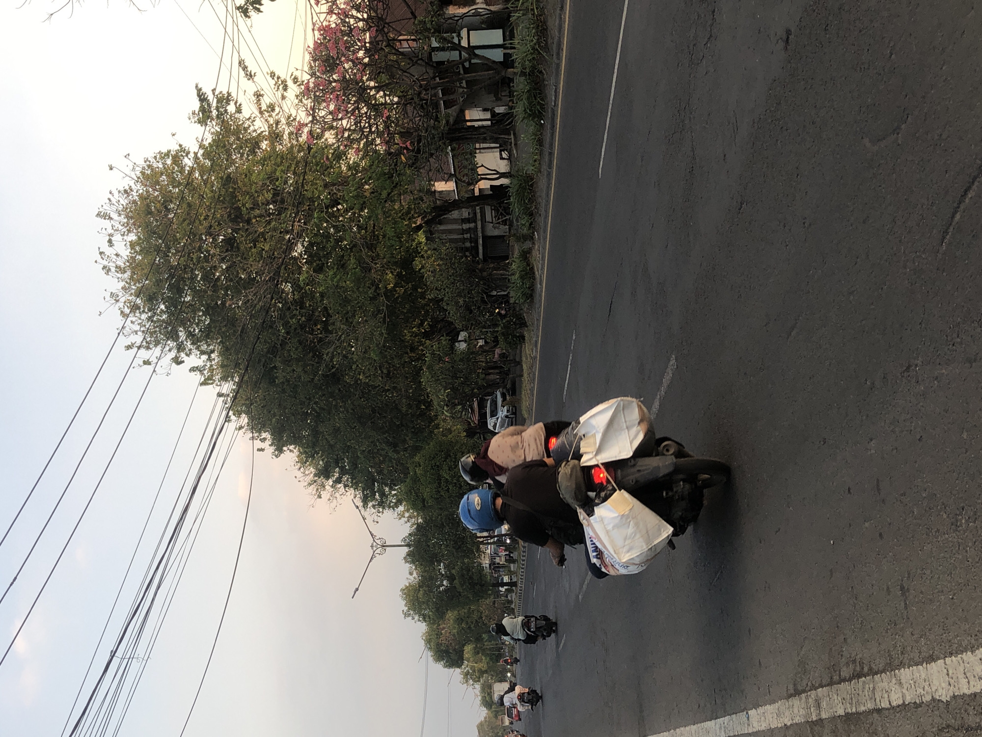 A street scene in Siring, Indonesia, shows a road with several motorcycles in motion. In the foreground, a motorcycle is ridden by two people. The rider wears a blue helmet and a black shirt, while the passenger wears a pinkish-brown shirt and a headscarf. The motorcycle has two large white bags strapped to the back. In the background, other motorcycles are visible, some with passengers. Trees line the side of the road, with some buildings partially obscured by foliage. Overhead power lines crisscross the sky. The lighting suggests it is either early morning or late afternoon.