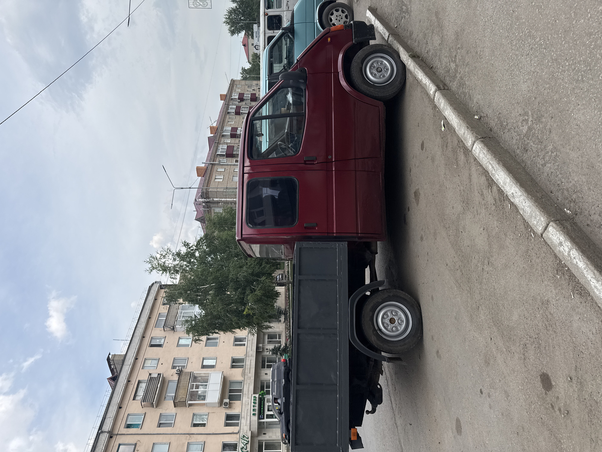 The image depicts a street scene in Kokshetau, Kazakhstan. A red Ford Transit van, modified with a flatbed truck body, is parked on the side of a paved road. The van is the primary subject, positioned in the center of the frame, facing to the right. Behind it, two other vans are partially visible: a light blue one to the immediate right, and a white one further back. The background features a multi-story apartment building with beige and pink facades and balconies, characteristic of Soviet-era architecture. A street with a concrete curb runs in front of the parked vehicles. A large green tree is situated between the apartment building and the van. The sky is overcast with clouds. No people are visible in the image, and the scene appears static, with no discernable actions taking place. The overall environment suggests a typical urban street in a post-Soviet city.