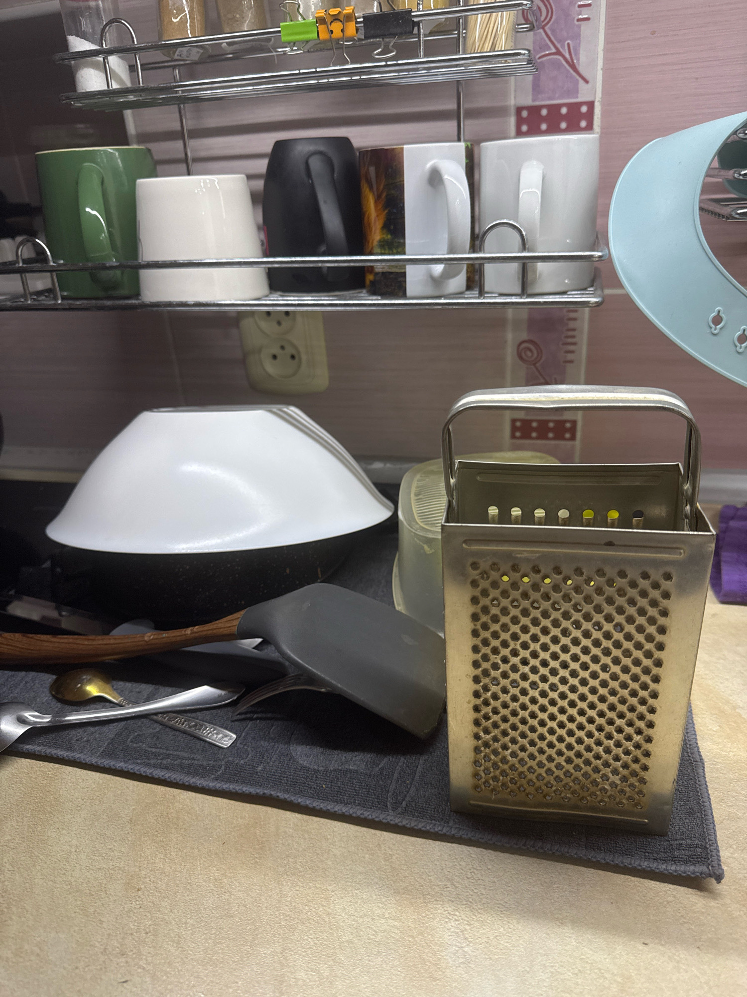 The image shows a kitchen counter with various items, including mugs on a shelf, a grater, a bowl, and utensils. The lighting suggests an indoor environment, likely a domestic kitchen. The specific location in the city of Hrodna, Belarus, cannot be determined from the visual information provided. The scene depicts everyday kitchen objects in a still life arrangement.