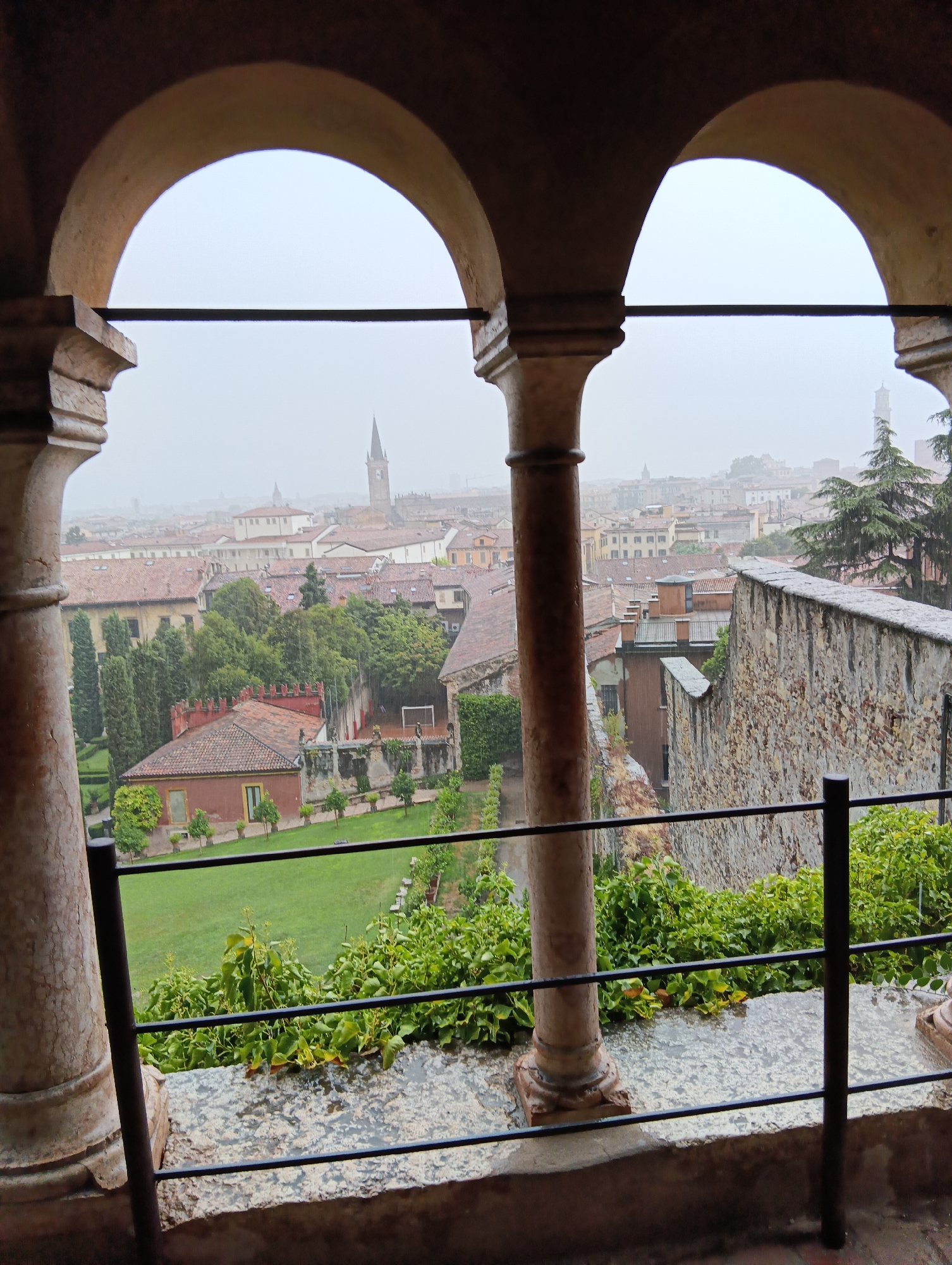 The image captures a view of the city of Verona, Italy, through an arched loggia. The foreground features two large, fluted columns and a stone balustrade, with a black metal railing running along the edge. Lush green foliage spills over the balustrade. Beyond the loggia, the cityscape unfolds under a hazy sky, suggesting overcast or foggy conditions. Numerous buildings with red-tiled roofs are visible, characteristic of Italian architecture. A tall clock tower or spire stands out against the horizon, and other architectural elements like chimneys and smaller towers are scattered throughout the urban landscape. The scene depicts an urban environment with a blend of historical architecture and natural elements.