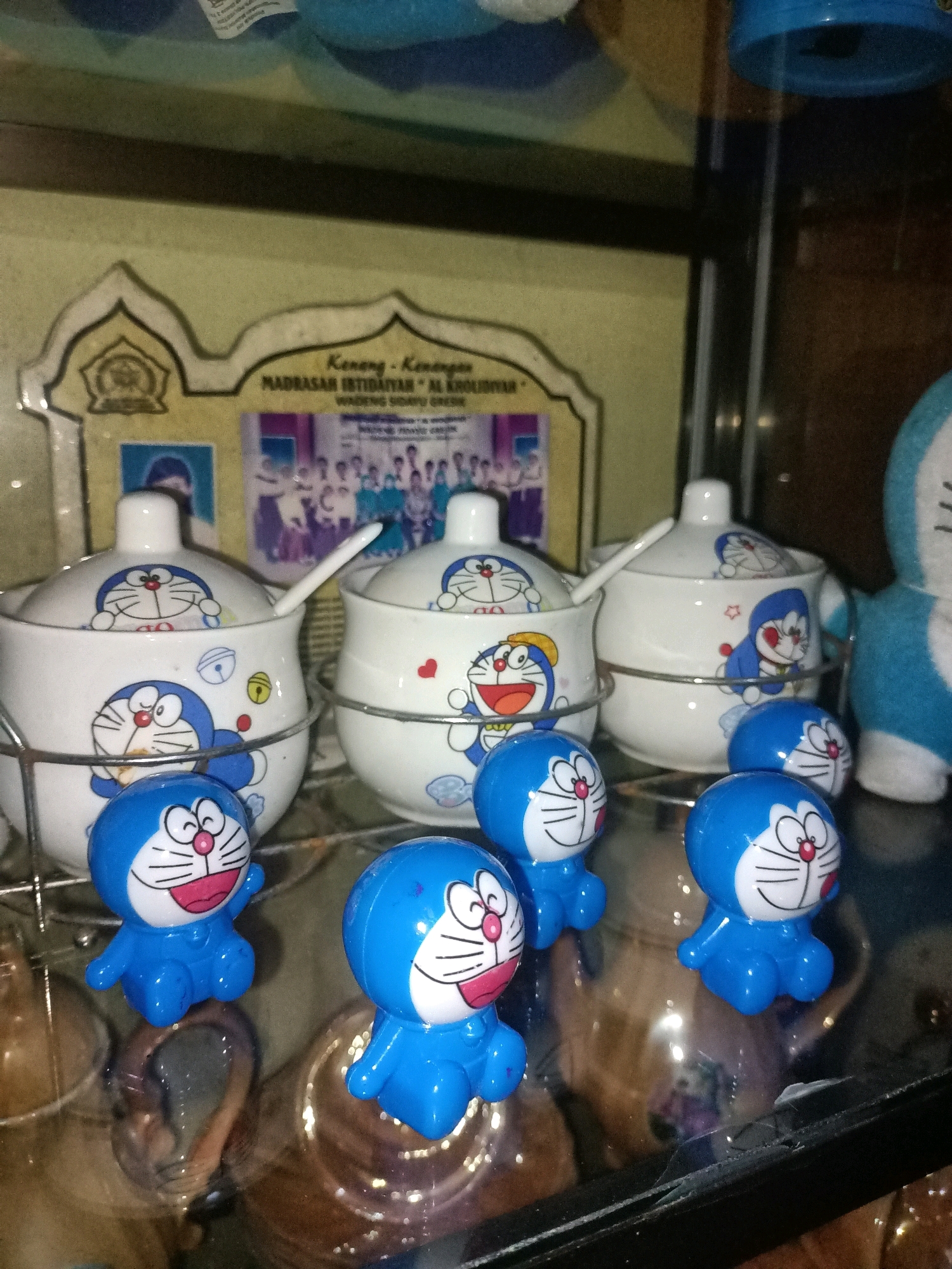 The image captures a close-up view of a shelf or display case, likely in a home or shop setting in Wadeng, Indonesia. The focus is on several Doraemon-themed items. In the foreground, there are three small, blue Doraemon figurines, each with a cheerful expression and a characteristic round face. Behind them, a collection of white ceramic containers, also adorned with Doraemon characters, are arranged on a metal rack. One of these containers has a small white spoon protruding from it. Further back, a framed certificate or diploma with text and images is visible, with the text "MADRASAH IBTIDAIYAH "AL KHOIRIYAH" WADENG SIDWU GRESIK" suggesting a connection to an educational institution in Wadeng, Gresik. To the right, a portion of a larger blue Doraemon plush toy is present. The overall lighting is dim, creating reflections on the glass surface of the display.