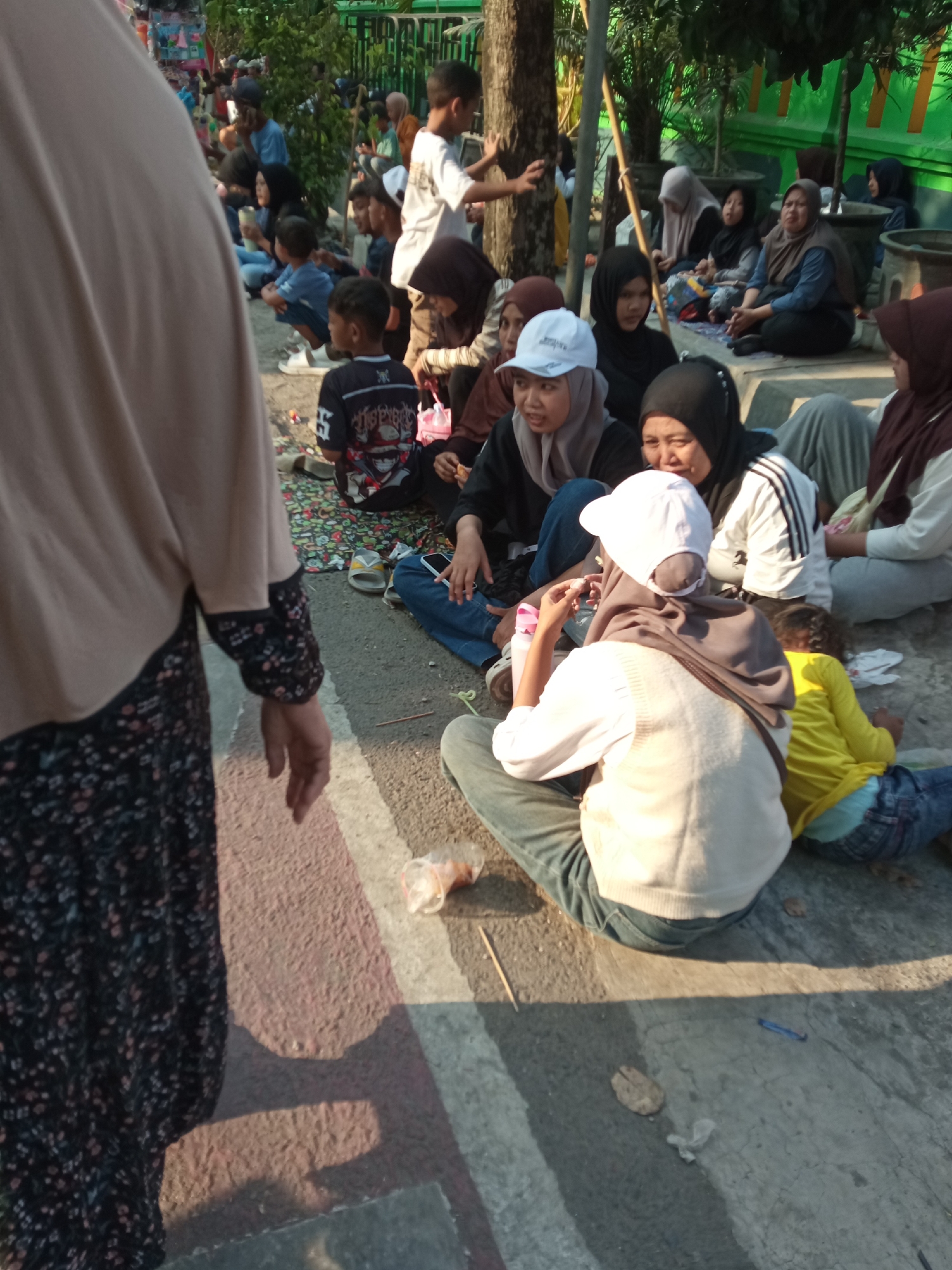 In the city of Balun, Indonesia, a group of people are gathered outdoors, seated on the ground and a low concrete ledge. The scene appears to be a street or public area, possibly near a park or event. The foreground is dominated by the back of a person wearing a tan hijab and a black and floral patterned dress, facing away from the camera.
To the right of this individual, a variety of people are seated on the ground. Several are wearing hijabs in various colors, including black, brown, and tan. Some individuals are children, including a boy in a black t-shirt with a graphic design and a younger child in a yellow shirt. One woman in a white hijab and a white baseball cap is looking to her left, holding a small object. Another woman in a black hijab is looking forward, while a woman in a brown hijab is looking to her right.
The ground is paved with asphalt and a light-colored concrete strip. There are some scattered objects on the ground, including a plastic cup, a leaf, and a blue pen. In the background, there are trees, a green fence, and more people, suggesting a larger gathering or public space. The lighting suggests it is daytime, with shadows cast on the ground.