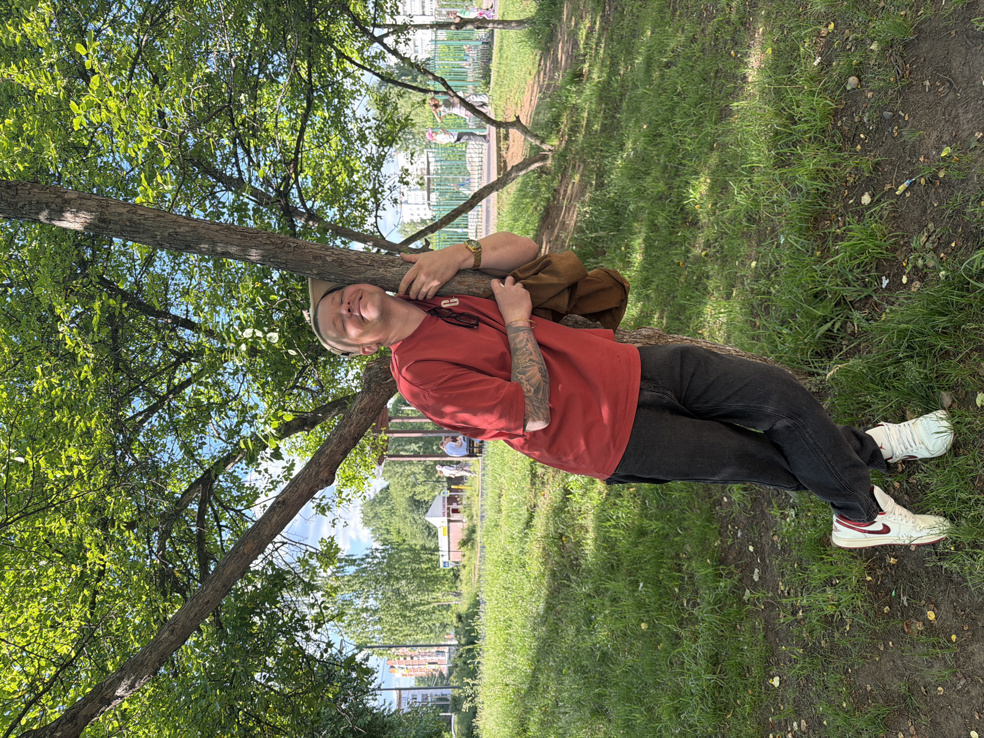 The image depicts an outdoor scene in a park or public green space within the city of Chaikovsky, Russia, during daylight hours. A fair-skinned individual, appearing male, with short light-colored hair and wearing a beige cap, is the primary subject. This person wears a red short-sleeved t-shirt, dark-colored jeans, and white high-top sneakers with red accents. Tattoos are visible on the left forearm and partially on the right upper arm. A gold-colored watch is on the left wrist and a gold-colored bracelet is on the right. Sunglasses are tucked into the t-shirt collar. The individual is leaning against the trunk of a deciduous tree, with eyes closed and a smile, conveying a contented expression. A brown fabric item, possibly a bag or jacket, is held in the left hand.
The immediate environment consists of green grass with patches of exposed soil and scattered fallen leaves or debris. Several trees with dense green foliage are present, casting dappled shadows on the ground.
In the background to the left, multi-story residential buildings are visible beyond a line of trees and a road. A small, light-colored building with a brown roof, resembling a kiosk, is also discernable in the mid-distance, with several indistinct figures standing nearby.
To the right in the background, a green metal fence separates the grassy area from a paved path. Beyond the fence, several other individuals are present, including two who appear to be children or young adults, one seemingly in motion. The sky, visible through the tree canopy, is blue with some white clouds. The scene is illuminated by bright natural sunlight, indicating midday or early afternoon.