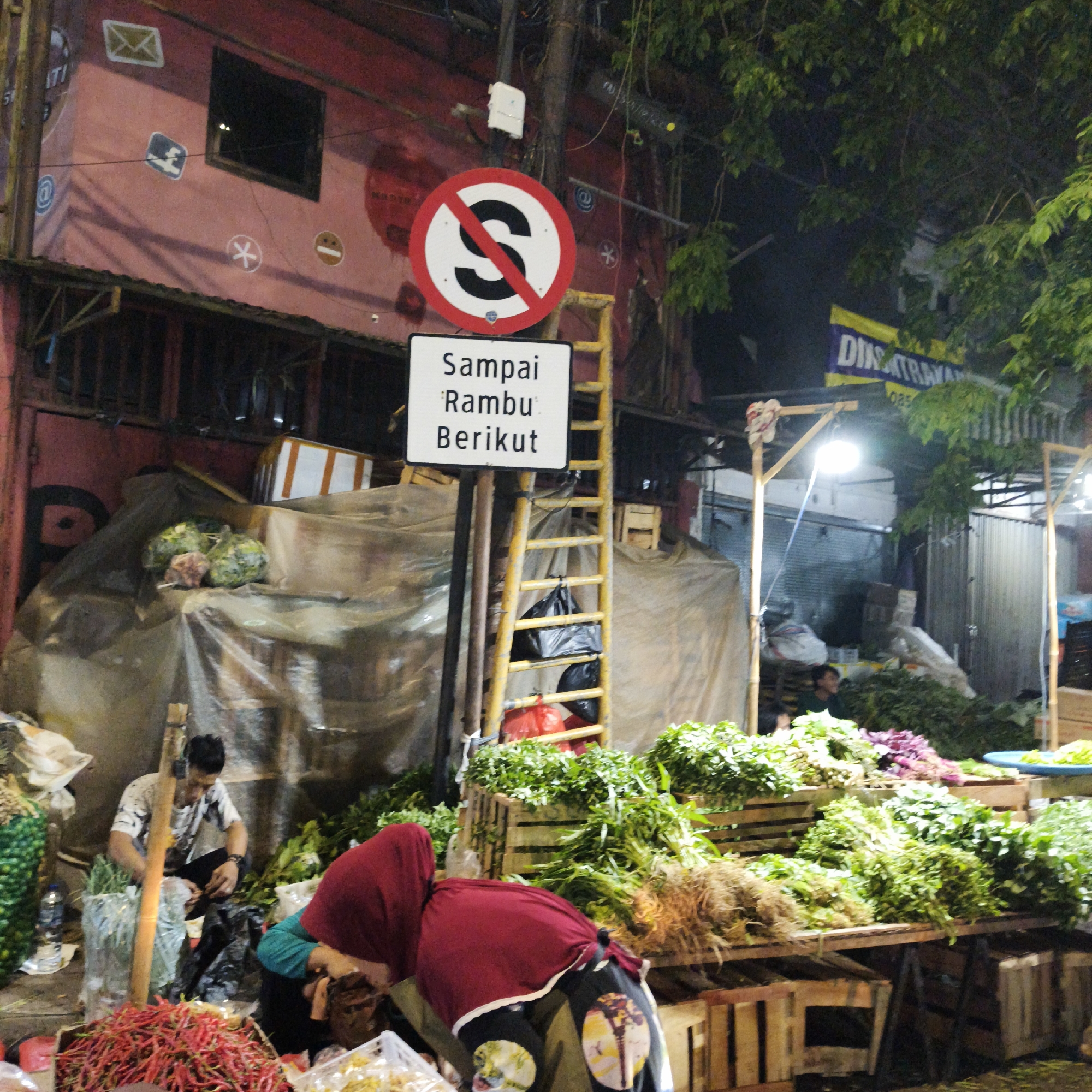 The image depicts an outdoor market scene at night, likely in South Jakarta, Indonesia, given the presence of Indonesian text on a sign. In the foreground, a stall is laden with a large pile of red chili peppers. Behind the chilies, two individuals are visible, one wearing a maroon headscarf and dark clothing, and another person partially obscured. A man in a patterned t-shirt is seated, appearing to be working with produce. The stall is covered by a translucent tarp.
In the midground, a yellow ladder leans against a pole next to a white sign with Indonesian text that reads "Sampai Ramu Berikut" which translates to "Until Next Sign." To the left of the ladder, various vegetables are displayed, some covered by tarps. A red and white circular sign with a black "S" crossed by a red diagonal line is prominently displayed on the pole above the text sign, indicating a prohibition. Further into the background, trees with dense foliage are visible, and a yellow banner with blue lettering is partially seen. The overall impression is a bustling, somewhat makeshift market environment illuminated by artificial light.