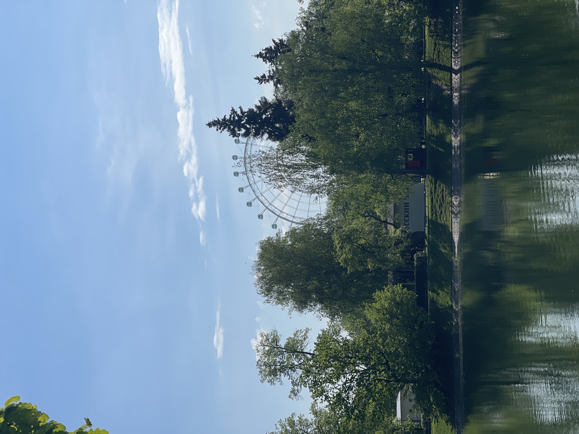 The image shows a large Ferris wheel partially obscured by trees, next to a body of water. The sky is blue with scattered clouds. The location appears to be in Gorky Park, Moscow, based on the style of the Ferris wheel and the presence of a sign in Russian. The water reflects the trees and part of the sky. A paved path runs along the edge of the water.