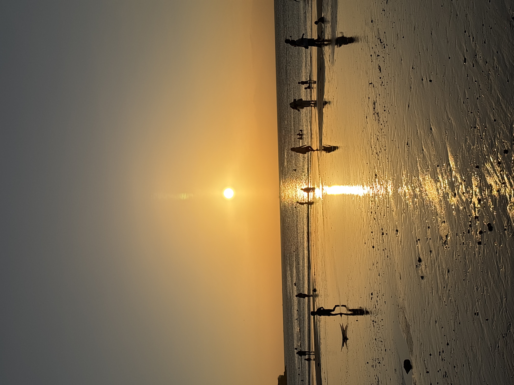 The image depicts a sunset scene at Kuta Beach in Kuta Selatan, Indonesia. Silhouettes of people are visible on the wet sand, some walking and others standing. A surfboard lies on the beach near a child who is running. The setting sun casts a golden reflection across the water and the wet sand, creating a shimmering effect. The ocean is calm, with gentle waves reaching the shore. The sky is a gradient of orange and yellow, transitioning to a darker hue towards the top of the frame. The overall atmosphere is serene and captures the beauty of a tropical beach at dusk.