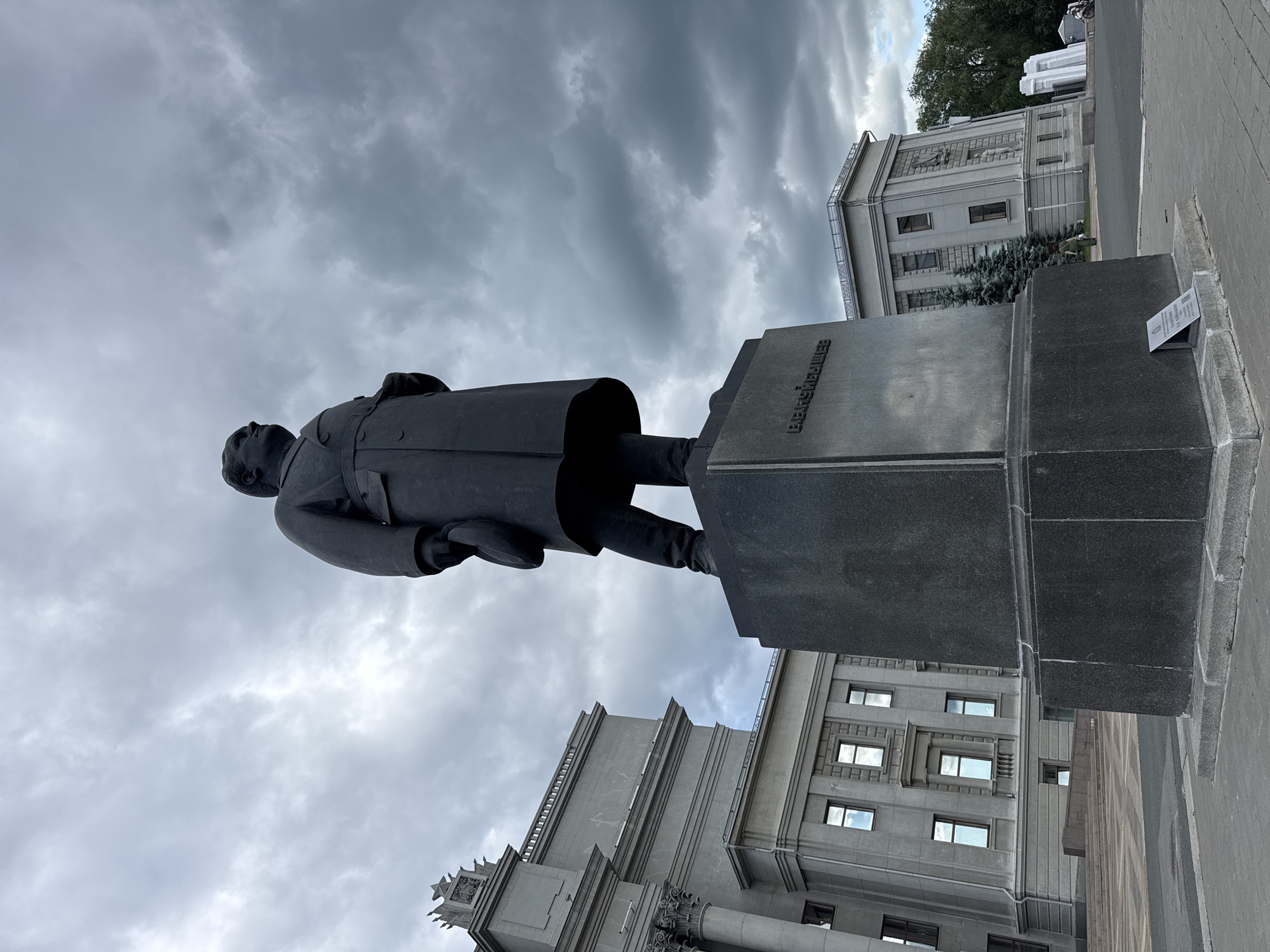 The image depicts a statue of a man in a long coat, holding a hat in his left hand. The statue stands on a tall, dark pedestal with an inscription in Cyrillic script. To the left of the statue is a large, gray building with classical architectural elements, including columns and ornate detailing. To the right, another gray building is visible, along with evergreen trees and a cloudy sky. The scene appears to be a public square or plaza, with paving stones in the foreground. The statue is located in Novokuybyshevsk, Russia.